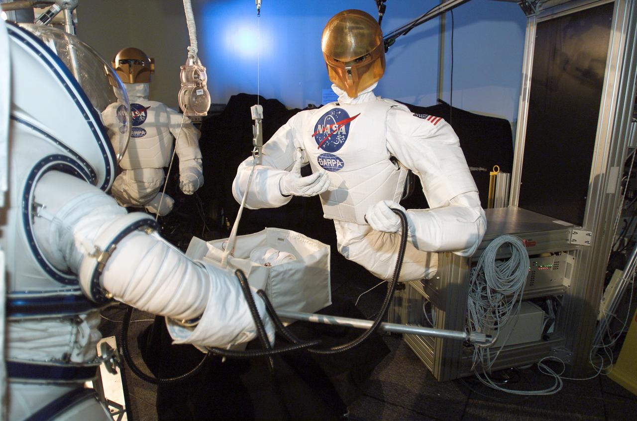 JSC2003-E-42622 (June 2003) --- Astronaut Nancy J. Currie (partially visible in foreground), wearing an advanced concept space suit, participates in a test at the Johnson Space Center to evaluate hand-in-hand work with robots. The Robonaut pictured is one of two that were used in the demonstration's task -- the assembly of an aluminum truss structure.