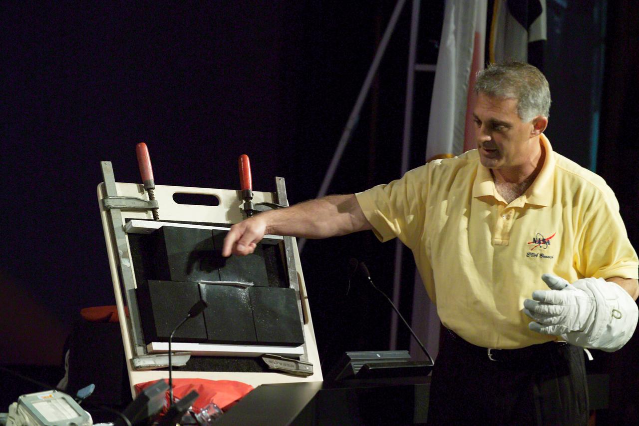 JSC2005-E-31523 (2 August 2005) --- Astronaut David A. Wolf points to a simulated piece of protruding fiber on a set-up of thermal tiles to demonstrate for a press conference audience possible techniques to extract the gap filler material on the underside of the Space Shuttle Discovery. The STS-114 crew will conduct a spacewalk on August 3 to remove two such pieces.