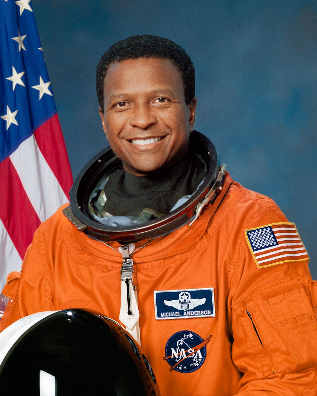 Official Portrait of Astronaut Mike Anderson (STS-107) in his orange launch and landing suit with an American flag in the background.