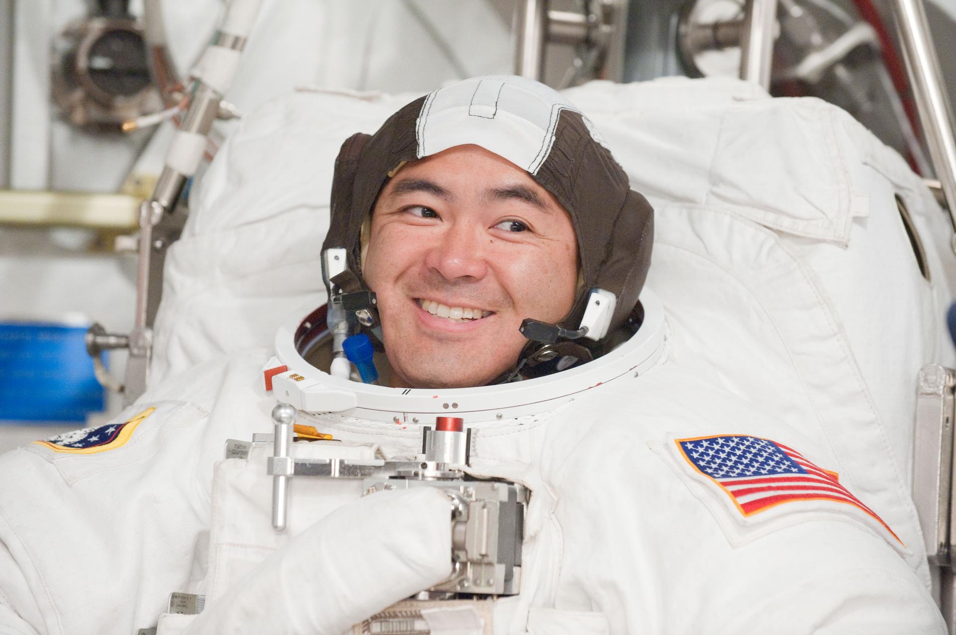 Astronaut wearing a spacesuit, smiling brightly while preparing for a mission. The suit features an American flag patch on the arm, and the astronaut is equipped with a helmet liner and communication gear. The background includes spacecraft hardware and equipment.