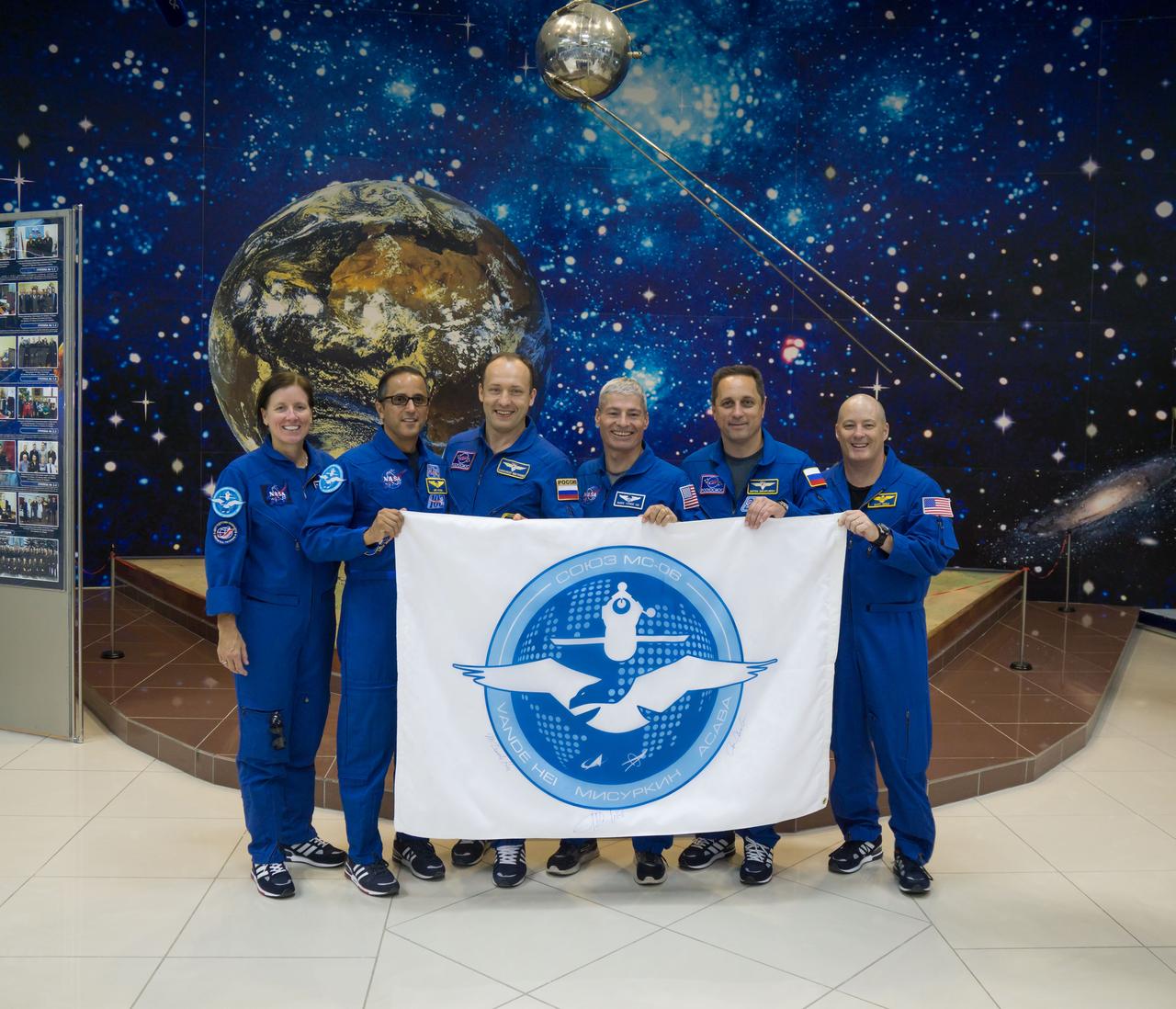 jsc2017e115133 (Sept. 7, 2017) --- In the Korolev Museum at the Baikonur Cosmodrome in Kazakhstan, the Expedition 53-54 prime and backup crewmembers display a flag bearing their Soyuz mission insignia Sept. 7 as part of traditional pre-launch activities. From left to right are Shannon Walker of NASA, Joe Acaba of NASA, Alexander Misurkin of Roscosmos, Mark Vande Hei of NASA, Anton Shkaplerov of Roscosmos and Scott Tingle of NASA. Acaba, Misurkin and Vande Hei will launch Sept. 13 on the Soyuz MS-06 spacecraft for a five and a half month mission on the International Space Station. Credit: NASA/Victor Zelentsov