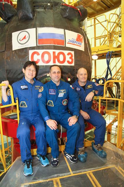 jsc2017e136448 - In the Integration Facility at the Baikonur Cosmodrome in Kazakhstan, Expedition 54-55 crewmembers Norishige Kanai of the Japan Aerospace Exploration Agency (JAXA, left), Anton Shkaplerov of the Russian Federal Space Agency (Roscosmos, center) and Scott Tingle of NASA (right) pose for pictures in front of their Soyuz MS-07 spacecraft Dec. 5 as part of their first fit check dress rehearsal activities. They will launch Dec. 17 in the Soyuz MS-07 vehicle for a five-month mission on the International Space Station...Courtesy of Roscosmos.