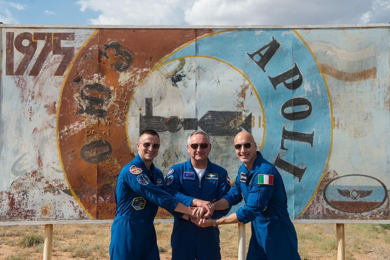 jsc2019e038379 - At the Baikonur Cosmodrome in Kazakhstan, Expedition 60 crewmembers Drew Morgan of NASA (left), Alexander Skvortsov of Roscosmos (center) and Luca Parmitano of the European Space Agency (right) pose for pictures July 5 in front of a mural bearing the insignia of the 1975 Apollo-Soyuz mission. They will launch July 20 on the 50th anniversary of humanity’s first landing on the moon during Apollo 11 on the Soyuz MS-13 spacecraft from the Baikonur Cosmodrome for a mission on the International Space Station...Andrey Shelepin/GCTC.