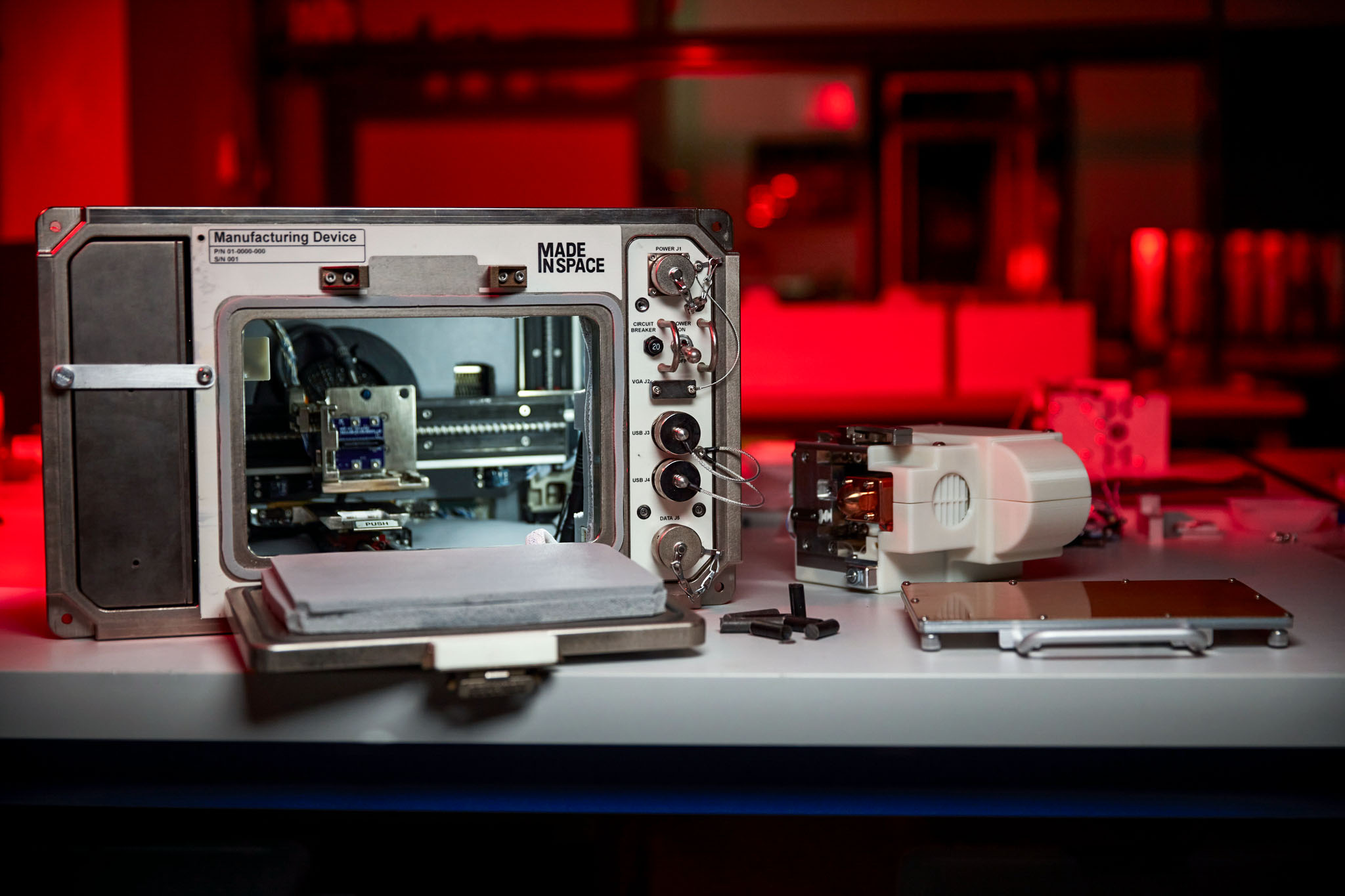 A metal box about the size of a microwave oven, sits on a gray work surface. A door on its front is open, revealing the metal printing mechanism inside. There is a white control panel to the right of the door. A laboratory in the background is blurred and bathed in red light.