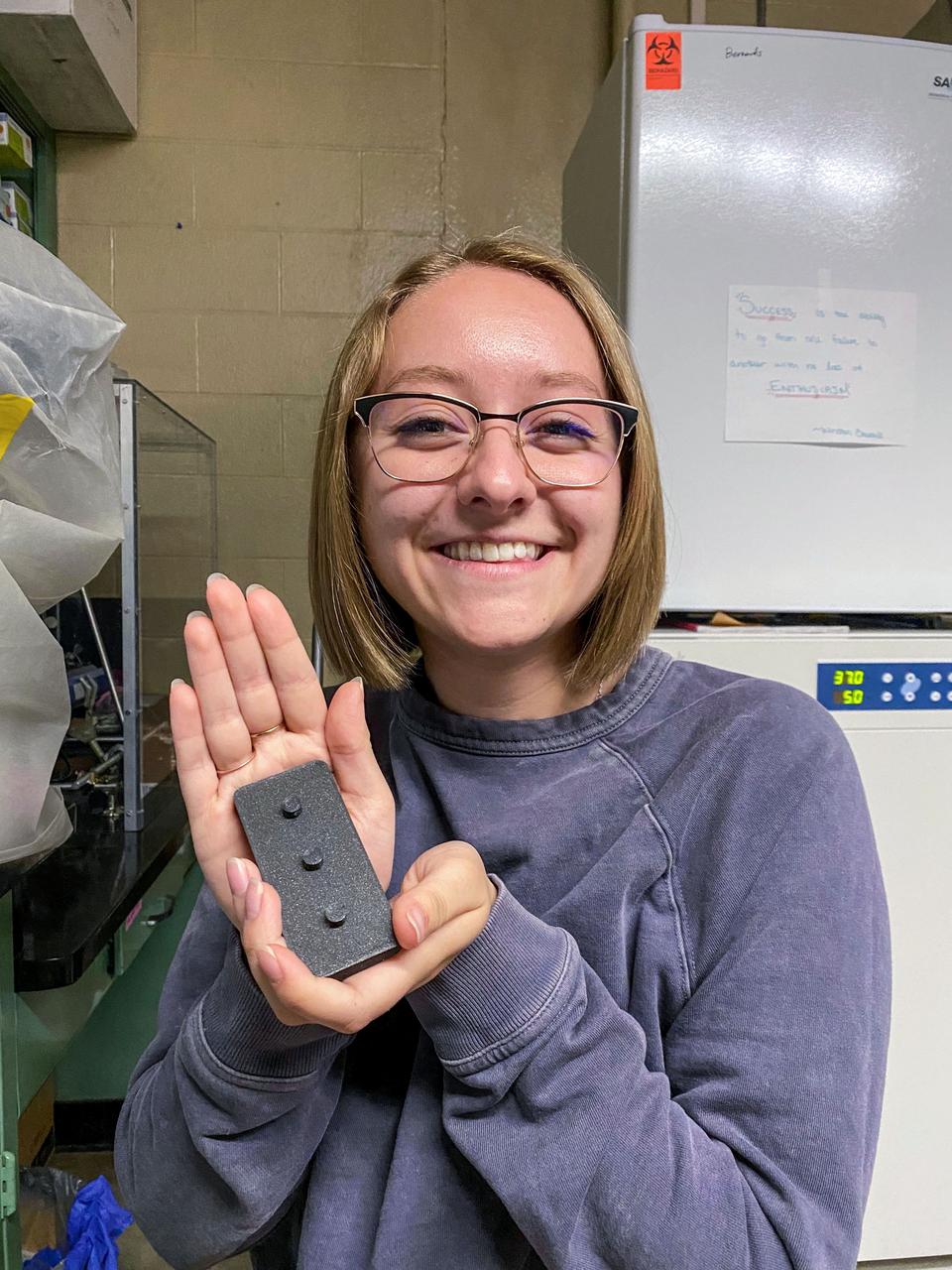 jsc2021e064551 (12/14/2021) --- Kaitlyn Harvey presents a 3D printed stand used to load and seal the bacteria introduction devices during assembly of the final experimental apparatus for the Determining the Efficacy of Bacteria Resistant Polymers in Microgravity (Bacteria Resistant Polymers in Space) investigation. Image courtesy of University of Idaho SPOCS Team.