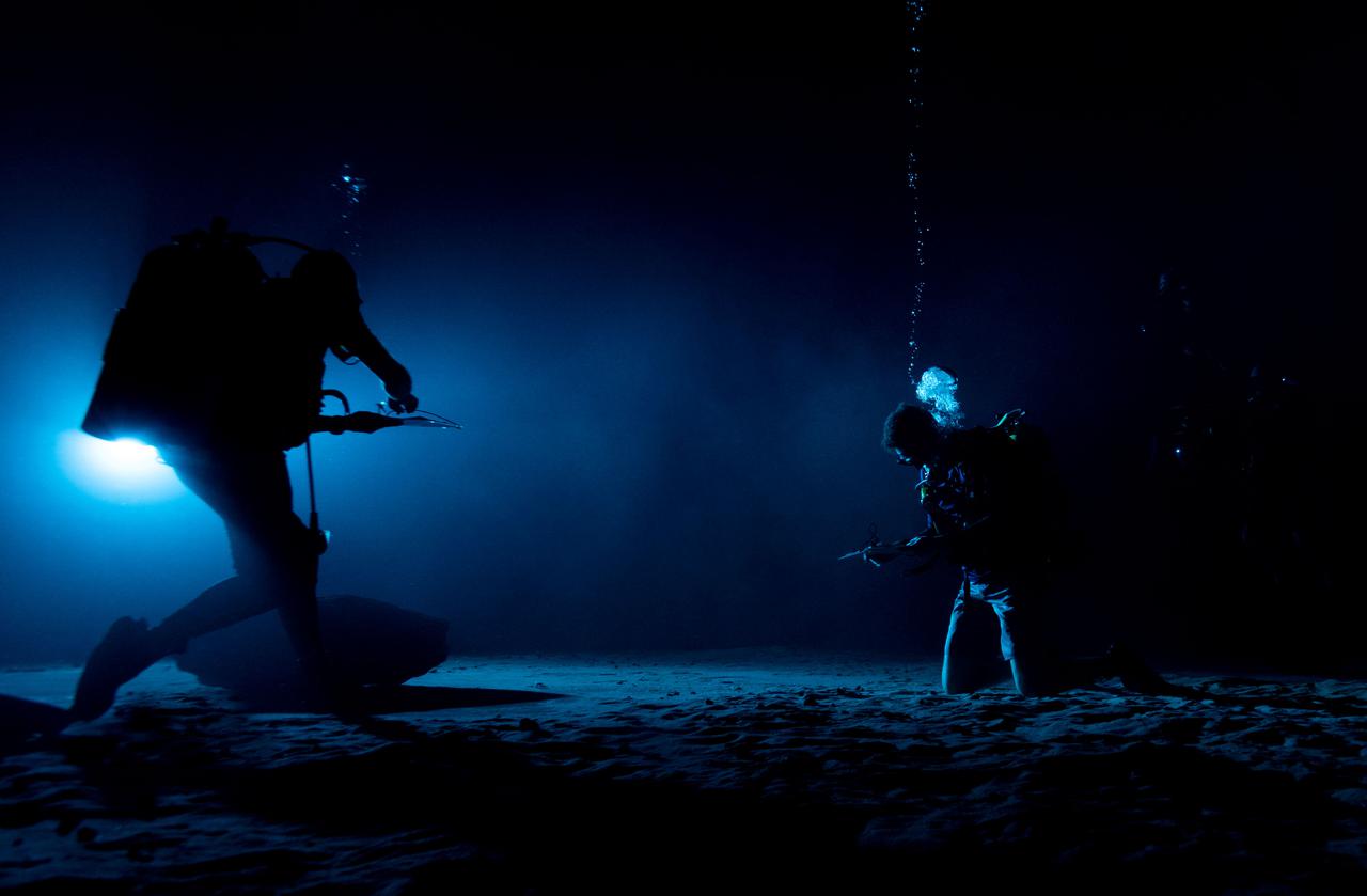 Test subjects performing subjective assessment of underwater lamp source during NBL Preliminary Lunar Lighting Evaluation.  Divers at the Neutral Buoyancy Laboratory (NBL) in Houston are setting the stage for future Moonwalk training by simulating lunar lighting conditions. At the Lunar South Pole, the Sun will remain no more than a few degrees above the horizon, resulting in extremely long and dark shadows. To prepare astronauts for these challenging lighting conditions, the team at the NBL has begun preliminary evaluations of lunar lighting solutions at the bottom of the 40-foot deep pool. This testing and evaluation involved turning off all the lights in the facility, installing black curtains on the pool walls to minimize reflections, and using a powerful underwater cinematic lamp, to get the conditions just right ahead of upcoming training for astronauts.