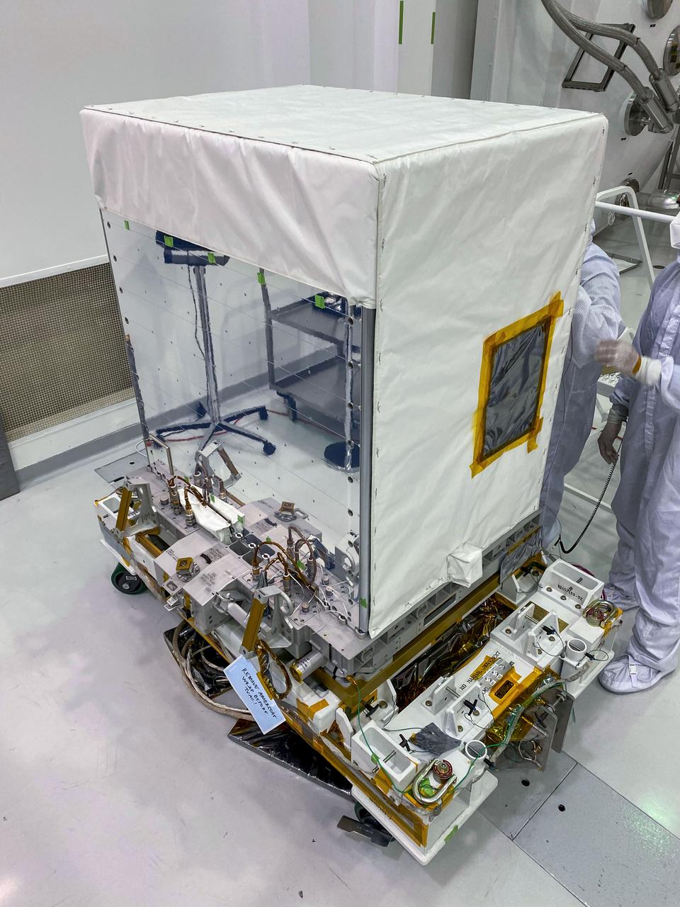 jsc2022e042615 (1/16/2022) --- The EMIT instrument after completion of all pre-launch environmental testing. The instrument is posed in front of one of the thermal-vacuum chambers that was used to qualify it for the hot and cold temperatures it will experience while operating on the exterior of the ISS. The reflective, mirror-like surface on the right is one of the instrument’s two radiator panels used to help cool the instrument electronics, cryocooler, and optics. The white cover is called “beta cloth,” which acts as a thermal blanket to keep the instrument within the desired operating temperature range. The dark gray portion surrounded by yellow tape is the telescope entrance. This covering prevents contaminants from being deposited on sensitive optical surfaces. Image courtesy of JPL.