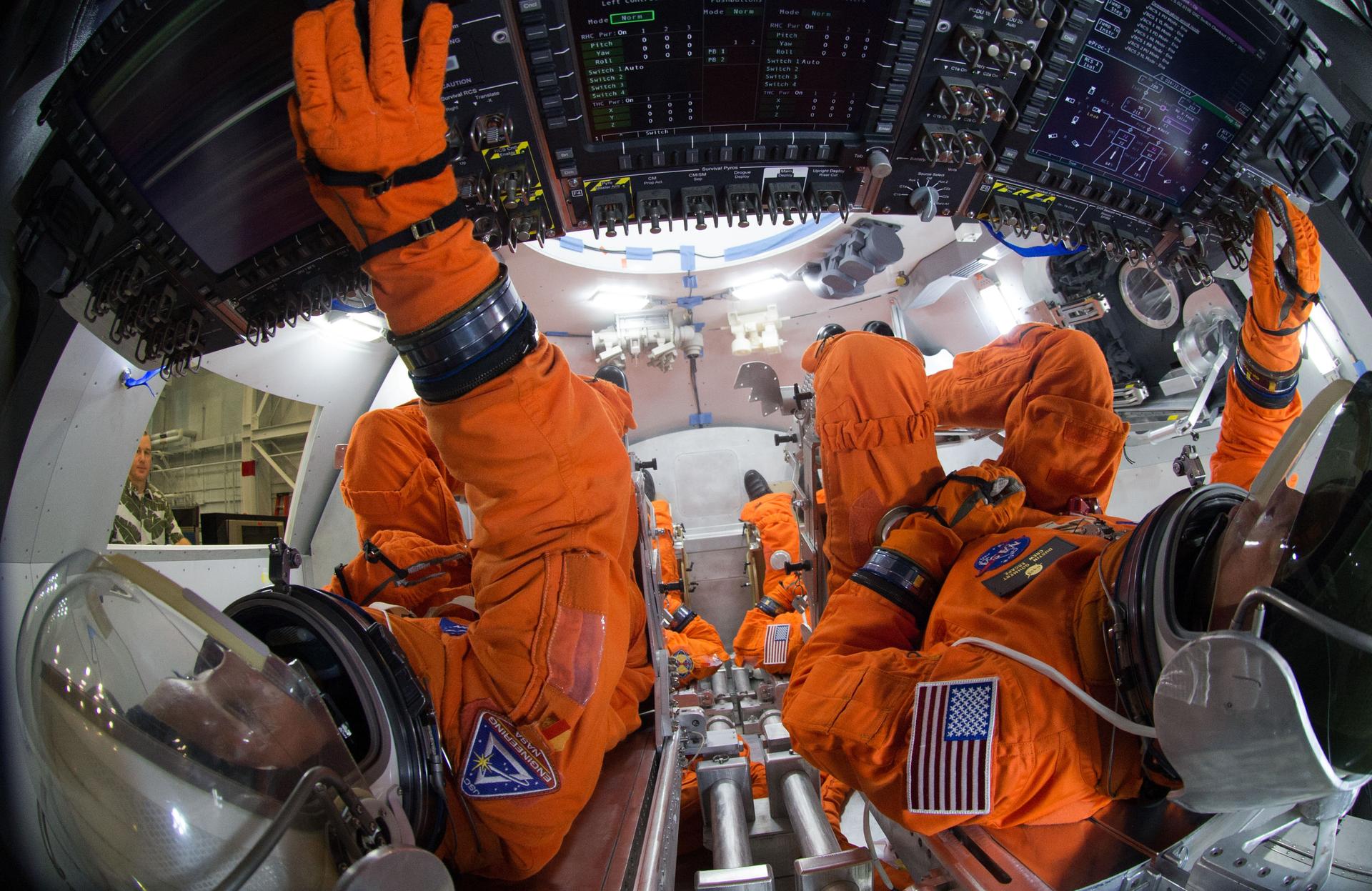 Engineers in orange spacesuits sit inside the Orion crew module mockup and touch the flat display panel.