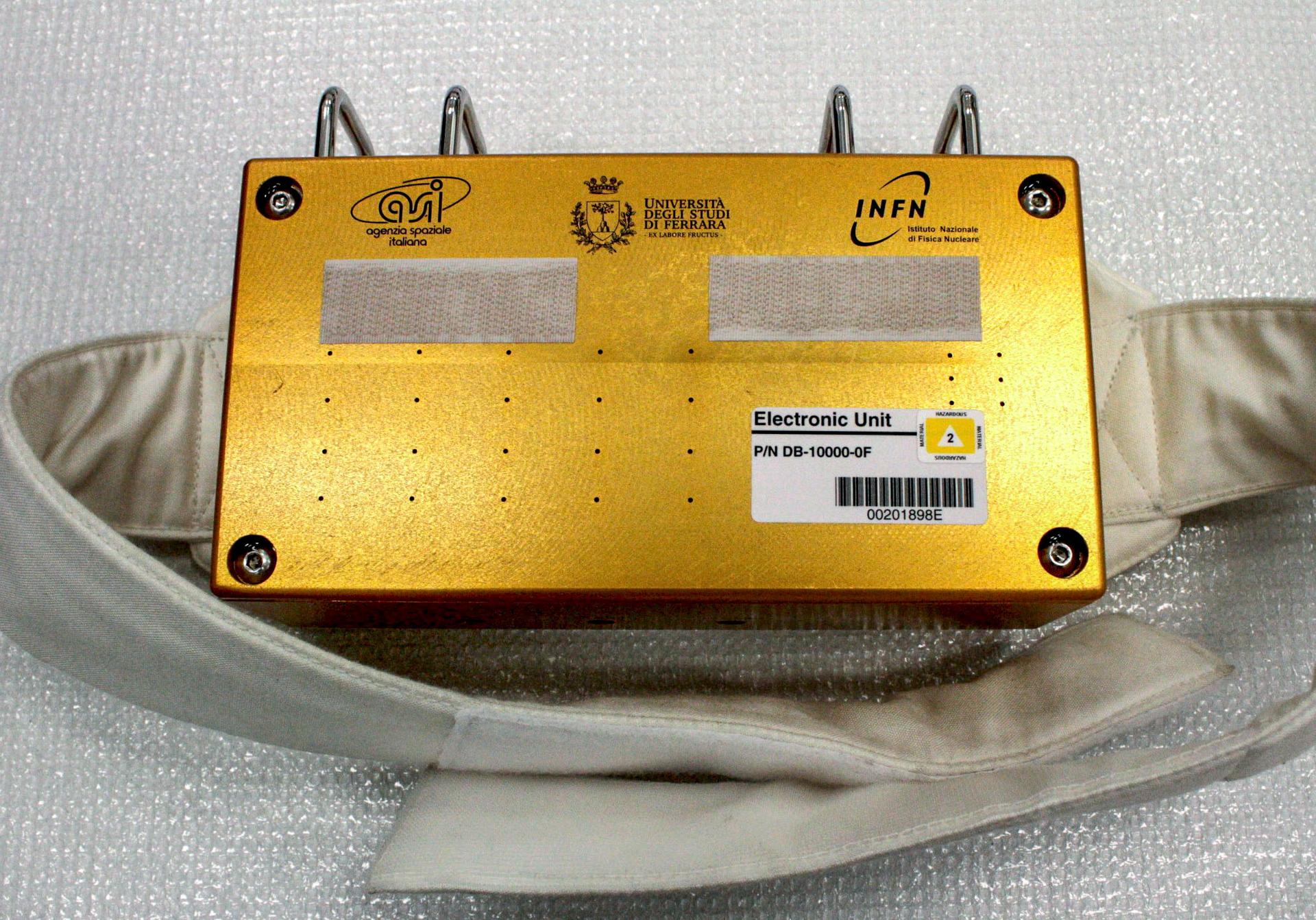 A shiny, gold rectangular box sits on a sheet of white plastic. The box has cloth straps coming out of each side and two strips of Velcro on its top along with a white barcode label and logos in Italian for the Italian Space Agency, University of Ferrara, and National Institute of Nuclear Physics.