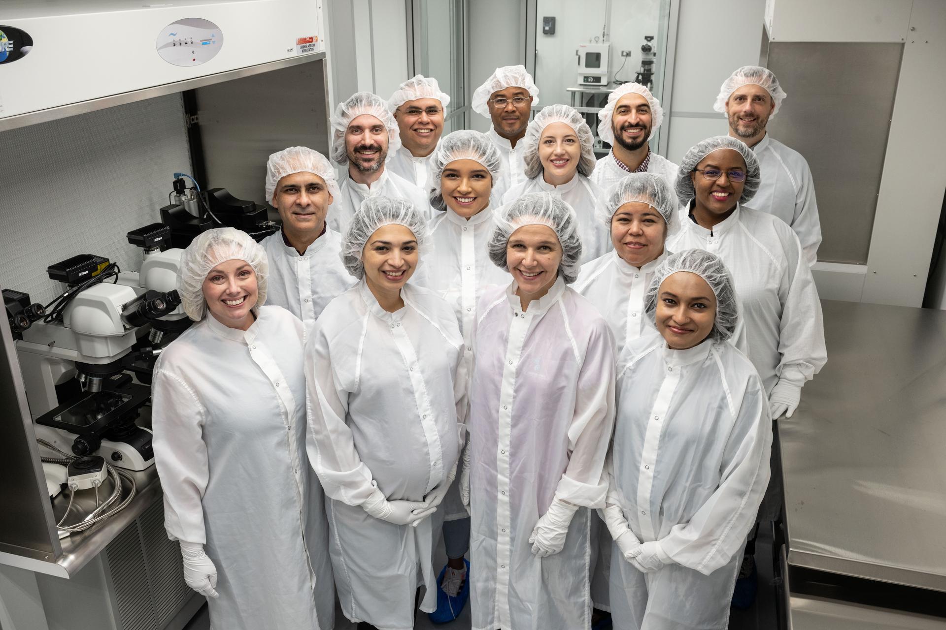 A group of people pose together in white lab coats and hair coverings. They are smiling inside a lab.
