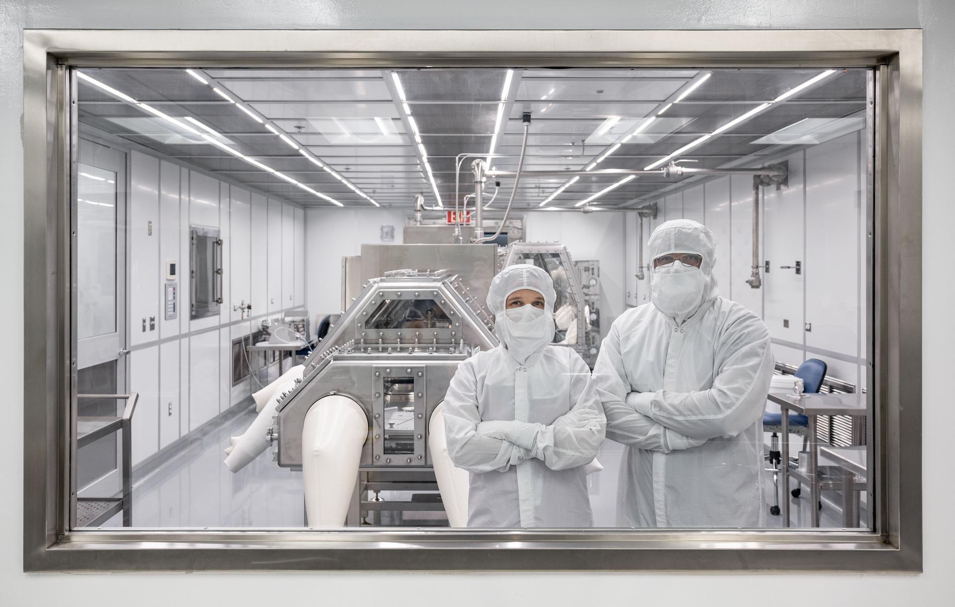 Two people in clean room suits pose on the other side of a window, into a clean room. The clean room contains a trapezoidal box with silver edges.