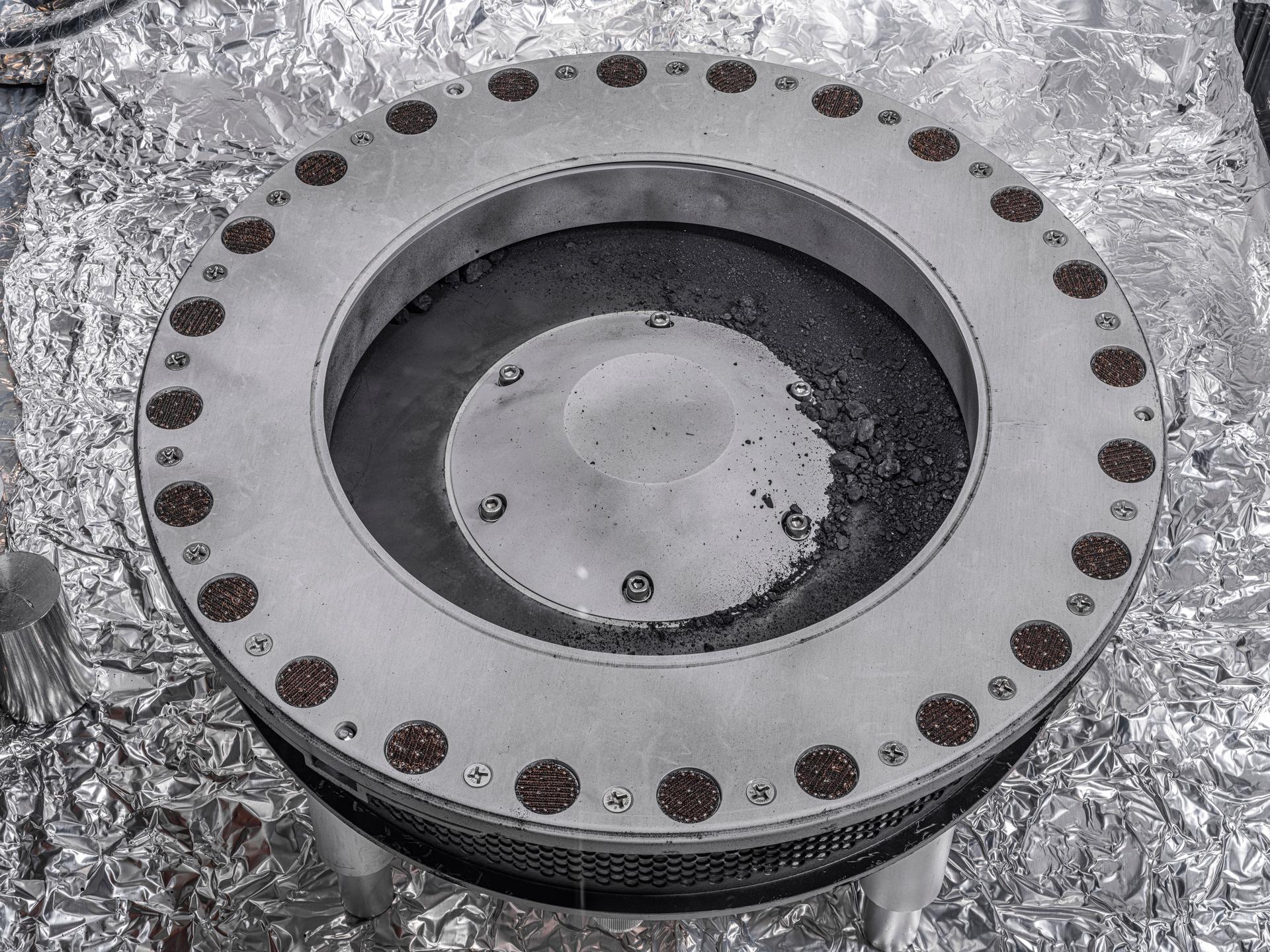 The TAGSAM head from the OSIRIS-REx spacecraft is circular metallic head, with concentric circles. The outer ring contains many small circles containing small particles of black dust. The inner circular segment contains small rocks.