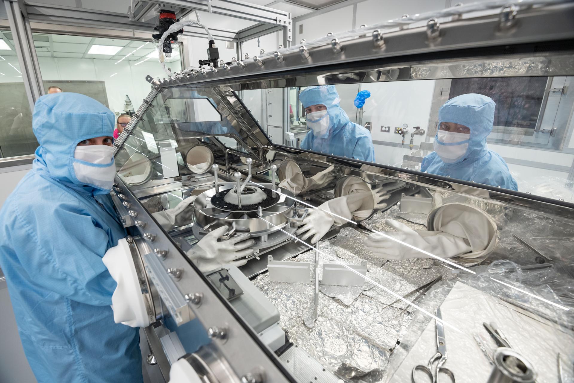 Three people in clean room hoods and masks on either side of a glove box containing the OSIRIS-REx TAGSAM capsule. The capsule is a squat silver cylinder, sitting on thin legs. The glove box is clear and has a trapezoidal top, so users can lean slightly over and see down into what they’re doing. Sets of white gloves extends into the box from both sides, so users can manipulate the sample capsule inside without exposing it to the outside world.