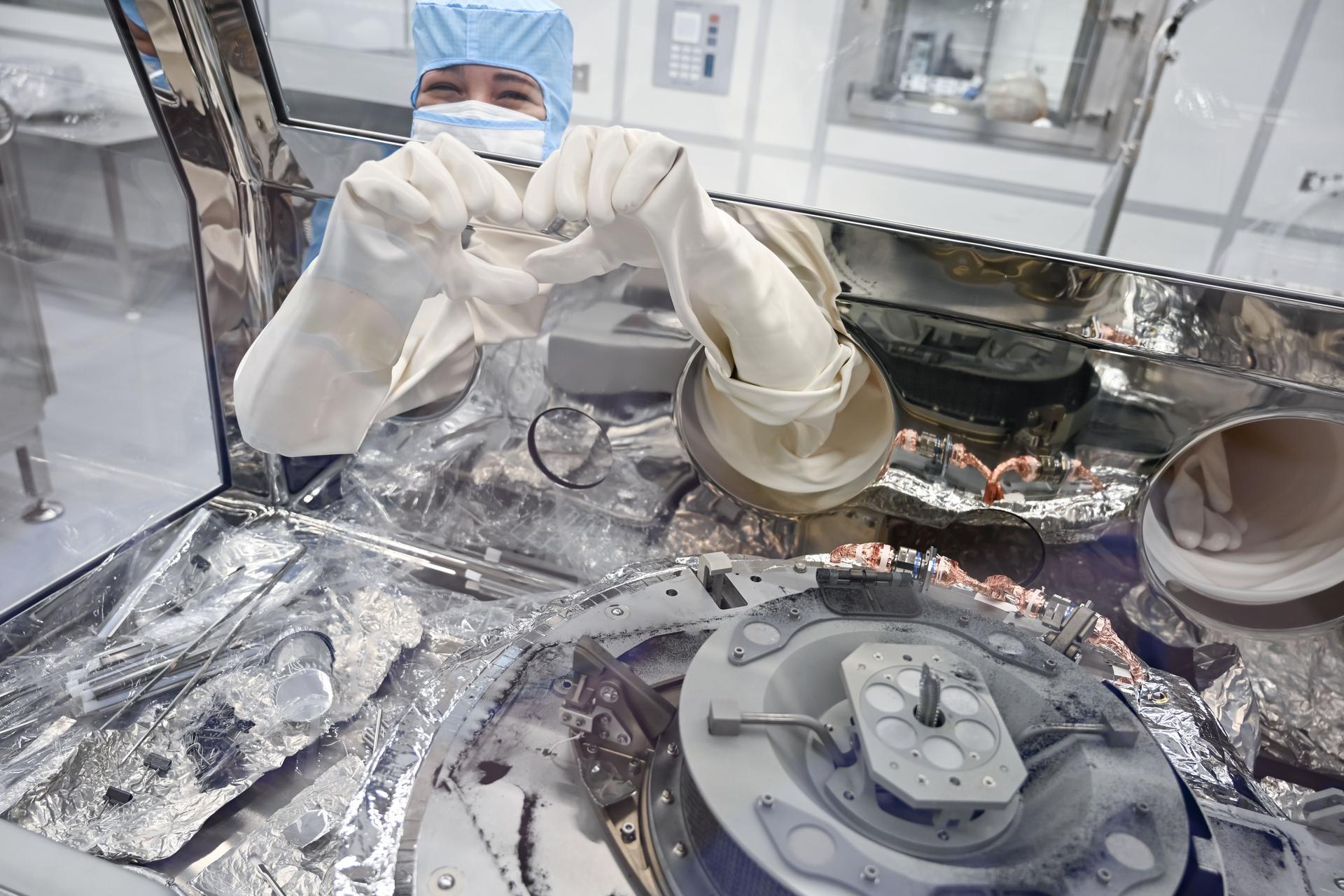 A person in a clean room hood and mask reaches into a glove box containing the OSIRIS-REx TAGSAM capsule. They use the white gloves extending into the glove box to make a hand heart. The capsule is a squat silver cylinder, sitting on thin legs. The glove box is clear and has a trapezoidal top, so users can lean slightly over and see down into what they’re doing.