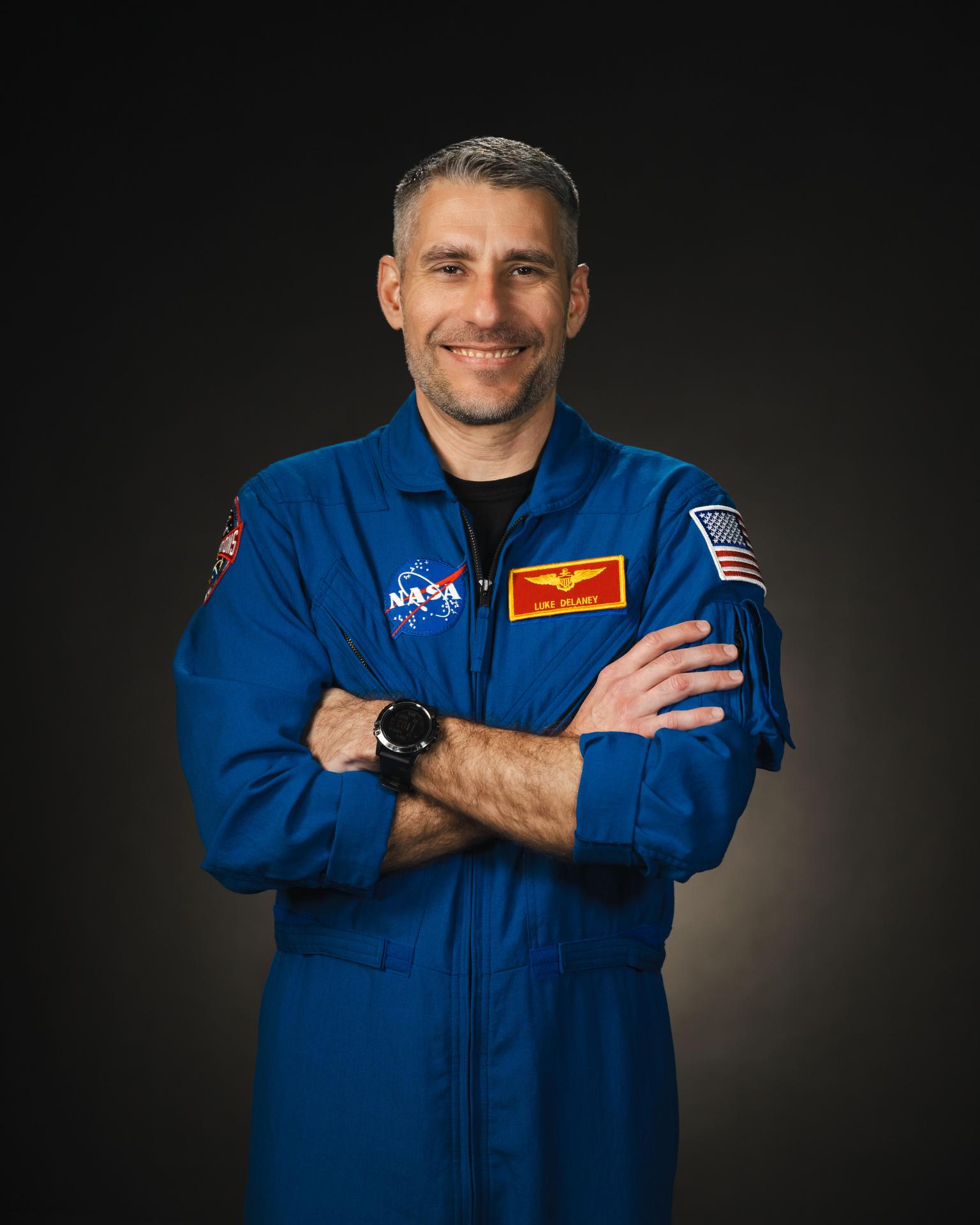 Image of an astronaut in a blue flight  suit standing with his arms crossed as he poses for a photograph. A NASA insignia patch is on his suit, along with his name in a red and yellow patch, along with an American flag patch on his arm.
