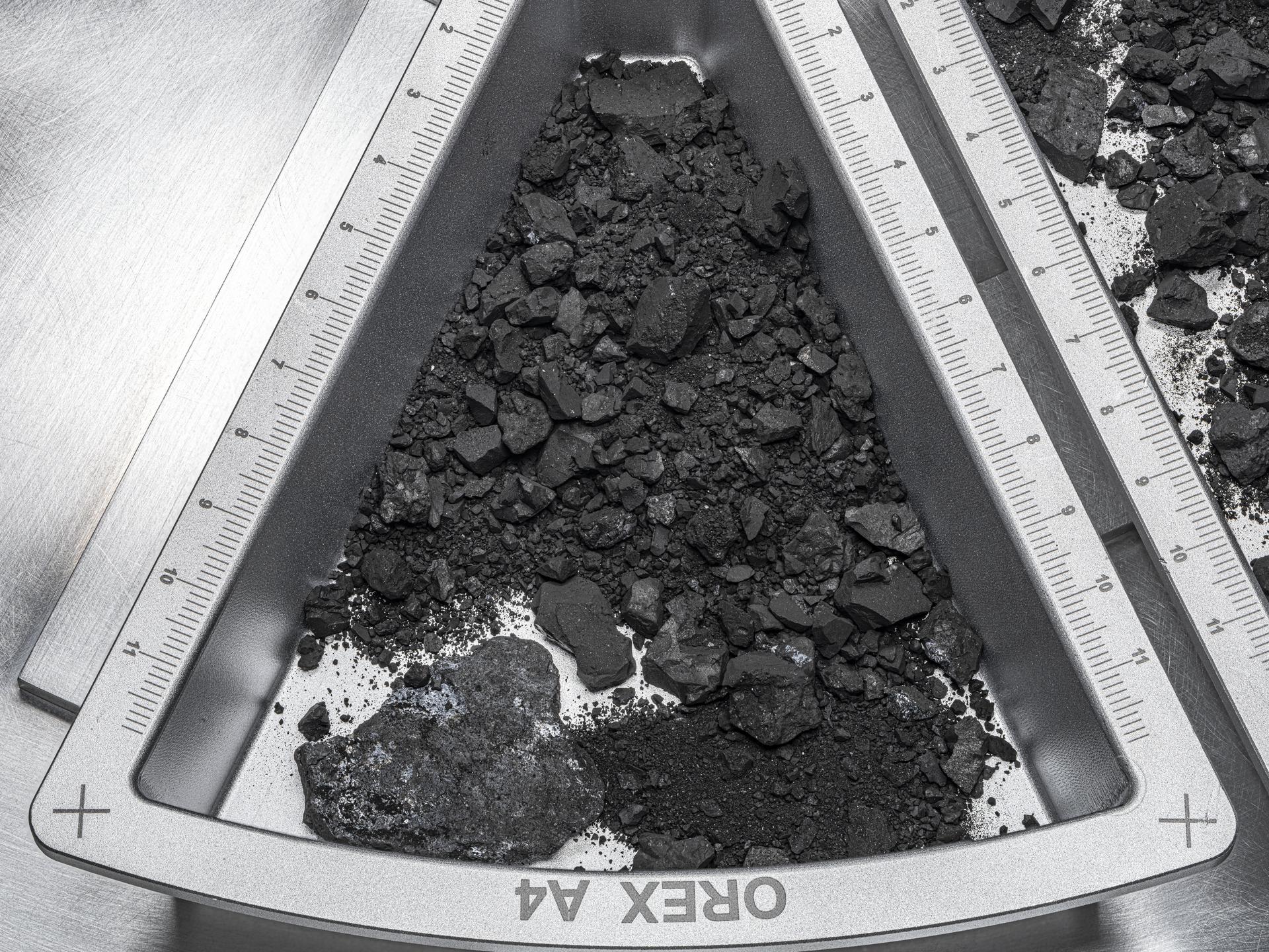 An image of a pizza-wedge-shaped, silver-colored tray with the pointy end at the top center of the image. In the tray is a pile of small, dark rocks and dust that fill most of the tray. Toward the left bottom corner of the tray is a larger rock that is slightly lighter in color and roughly shaped like a heart on its side.