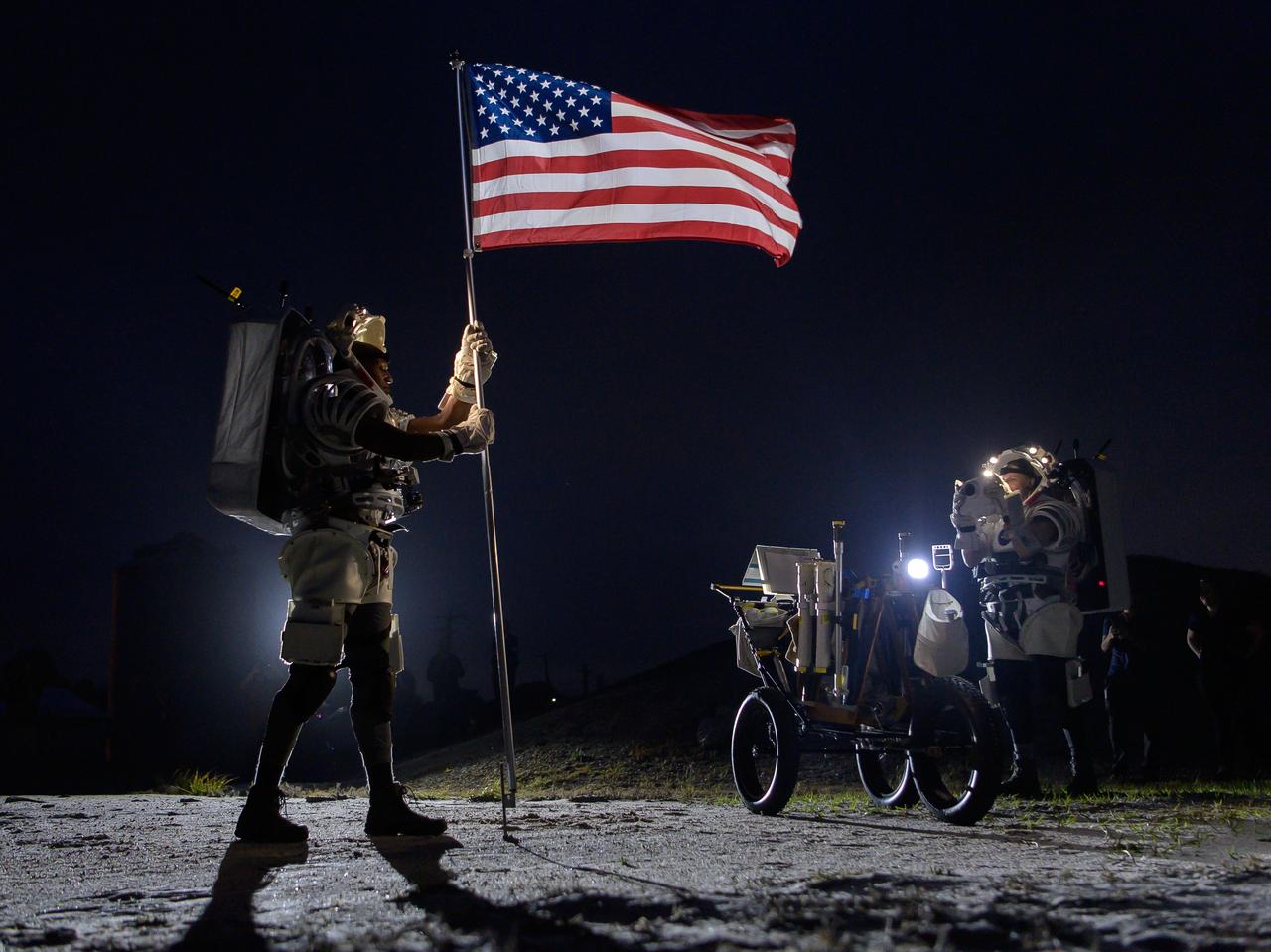 jsc2024e031840 --- NASA astronaut Kate Rubins (right) takes a photo of NASA astronaut Andre Douglas (left) as he raises an American flag during a simulated moonwalk in a rock yard at NASA’s Johnson Space Center. Credit: NASA/Josh Valcarcel