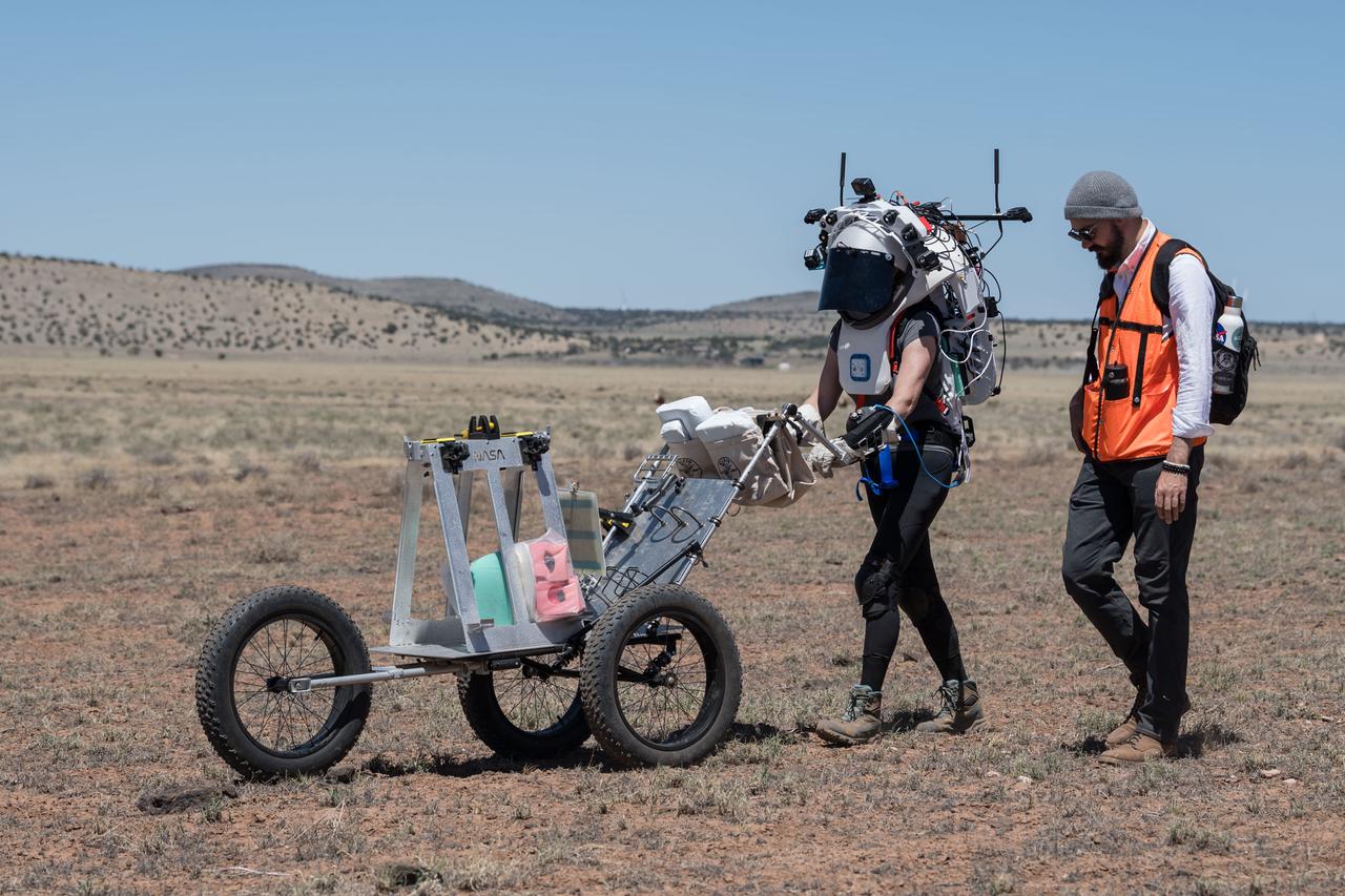 NASA astronaut Kate Rubins pushes a cart through the lunar-like landscape while wearing the Joint AR (Joint Augmented Reality Visual Informatics System) display during an advanced technology run in the San Francisco Volcanic Field in Northern Arizona on May 19, 2024. The suit display features include navigation, photo capture, graphical format of consumables, procedure viewing, mission control updates, and other augmented reality cues and graphics. The team successfully tested navigation displays using data from four different data streams: GPS (Global Positioning System)/IMU (Inertial Measurement Unit), camera/IMU, LiDAR (Light Detection and Ranging), and static maps. Technology like this may be used for future Artemis missions to augment mission control communication and help guide crew back to the lunar lander. Credit: NASA/Josh Valcarcel