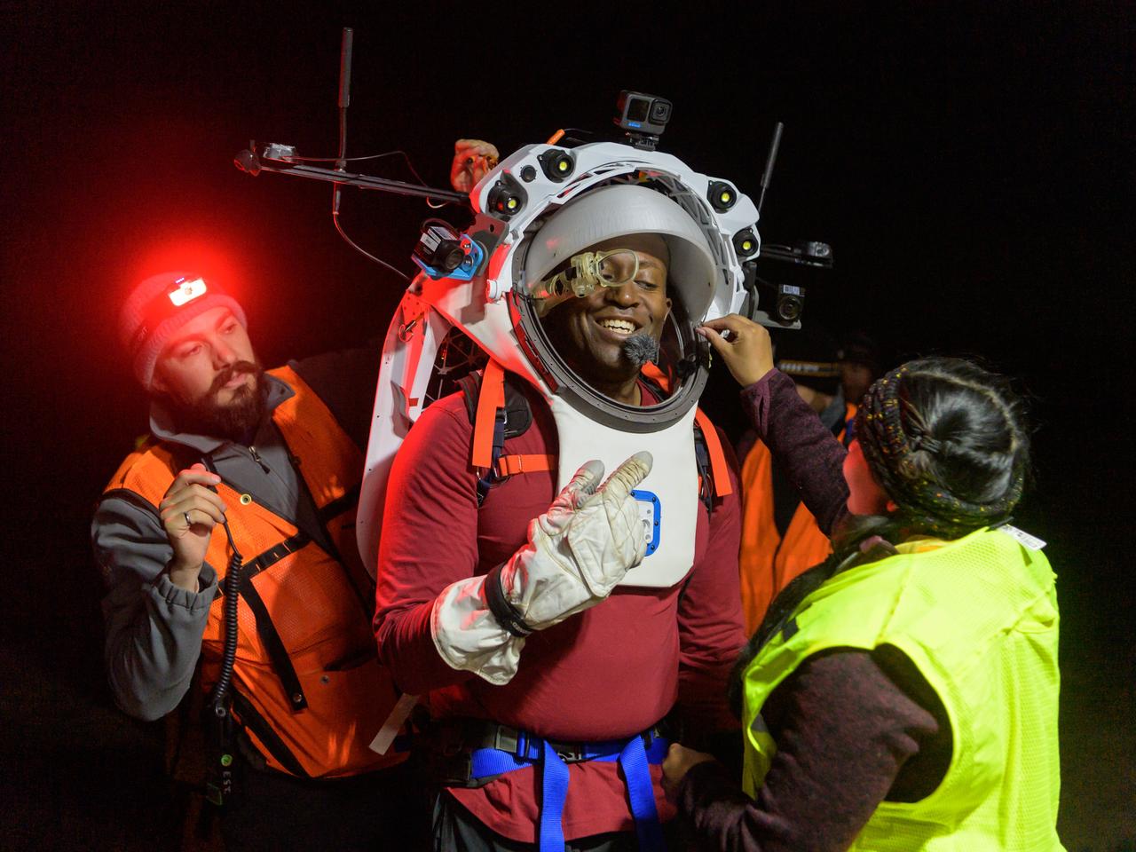 Engineers help NASA astronaut Andre Douglas adjust the Joint AR (Joint Augmented Reality Visual Informatics System) display he’s wearing during a nighttime advanced technology run in the San Francisco Volcanic Field in Northern Arizona on May 21, 2024. The suit display features include navigation, photo capture, graphical format of consumables, procedure viewing, mission control updates, and other augmented reality cues and graphics. The team successfully tested navigation displays using data from four different data streams: GPS (Global Positioning System)/IMU (Inertial Measurement Unit), camera/IMU, LiDAR (Light Detection and Ranging), and static maps. Technology like this may be used for future Artemis missions to augment mission control communication and help guide crew back to the lunar lander. Credit: NASA/Josh Valcarcel