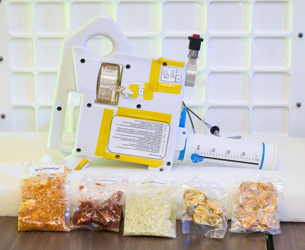 jsc2024e040685 -- A prototype of the Mini Potable Water Dispenser, currently in development at NASA’s Marshall Space Flight Center, is displayed alongside various food pouches during a demonstration at NASA’s Johnson Space Center. Photo Credit:  NASA/David DeHoyos