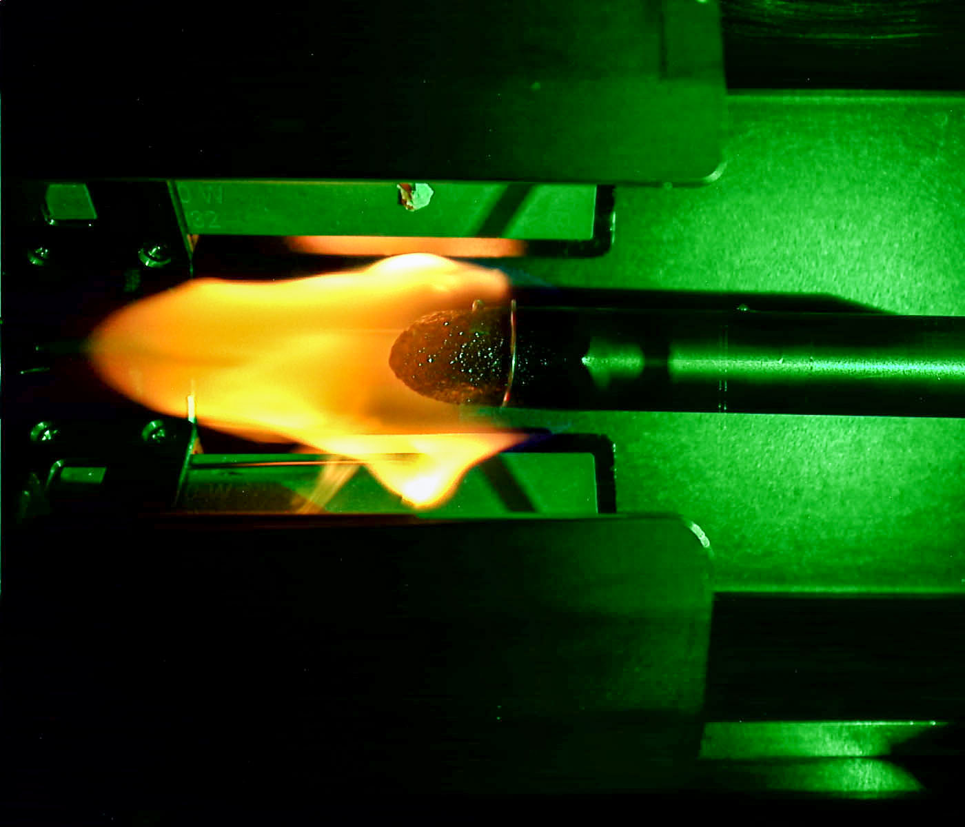 A plexiglass rod extending horizontally from right to left has a bright yellow and orange flame on its tip. The rod is inside a metal structure bathed in green light.