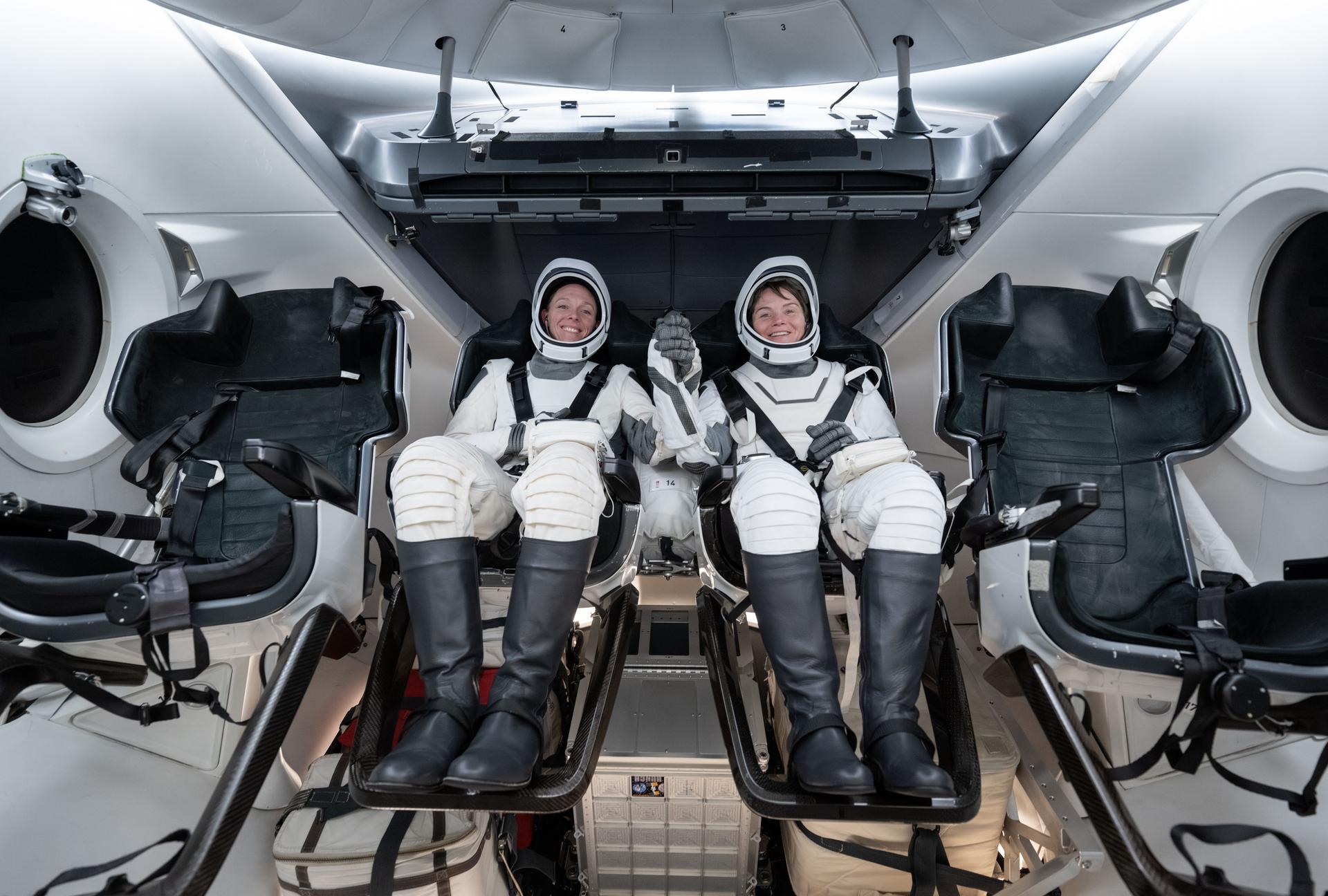 NASA astronauts (from left) Nichole Ayers and Anne McClain, NASA's SpaceX Crew-10 mission pilot and commander respectively, smile and grab hands during a simulation inside a mockup of a Dragon cockpit at the company's facilities in Hawthorne, California.