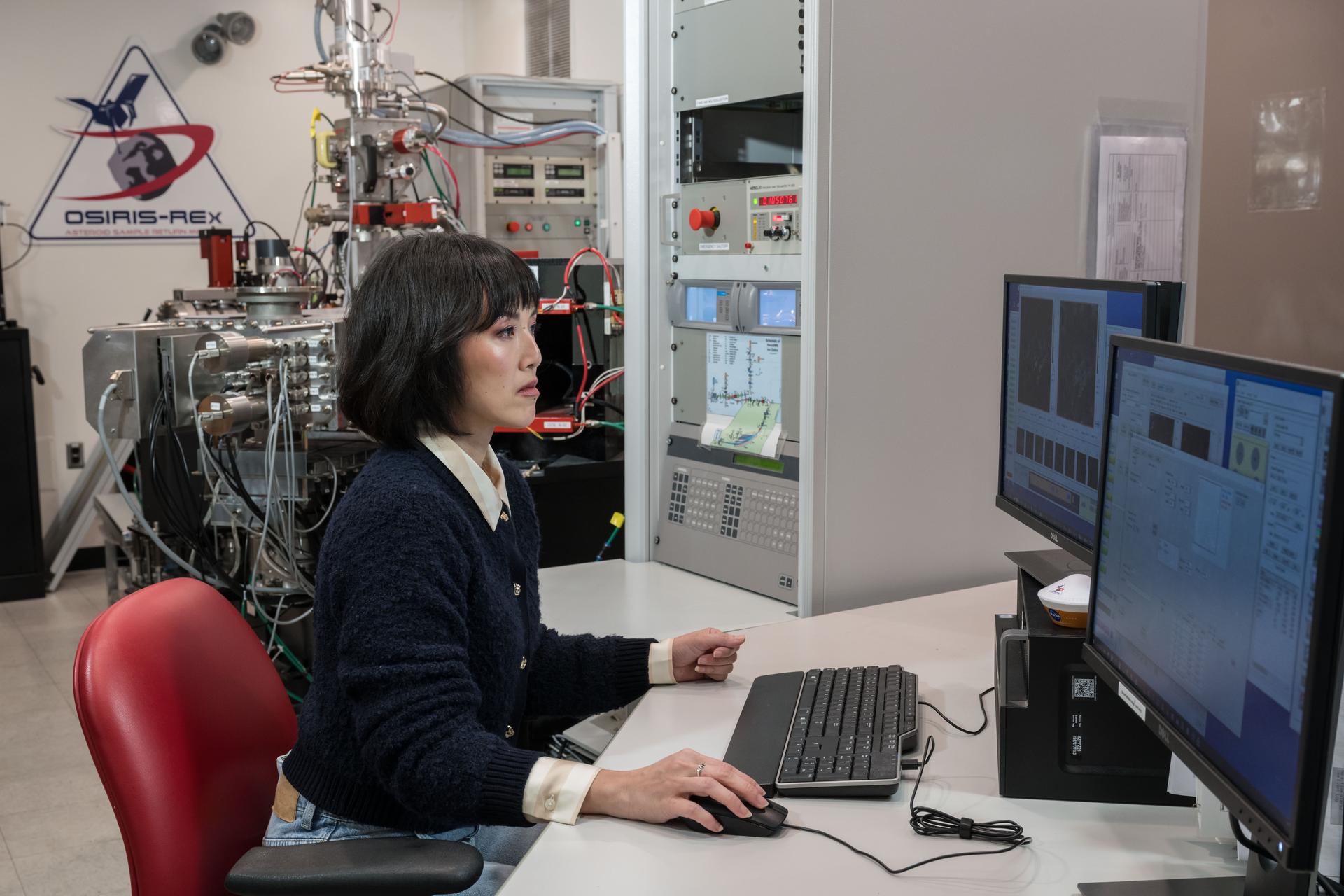 A woman sits in front of a computer with two monitors on a desk. Scientific instruments and the OSIRIS-REx logo are visible behind her.