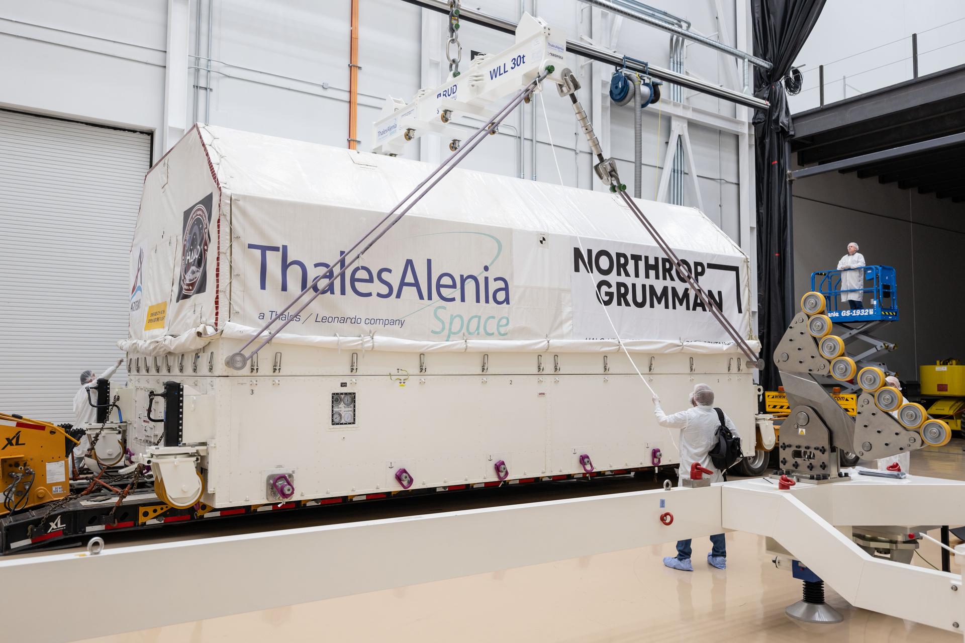 Personnel guide the lift of HALO’s transport container inside Northrop Grumman’s facility in Gilbert, Arizona. The white container, labeled with Thales Alenia Space, Northrop Grumman, Artemis, and HALO logos, is secured by straps and suspended by an overhead crane. A technician on a lift observes the operation from above as the container is positioned for unloading.