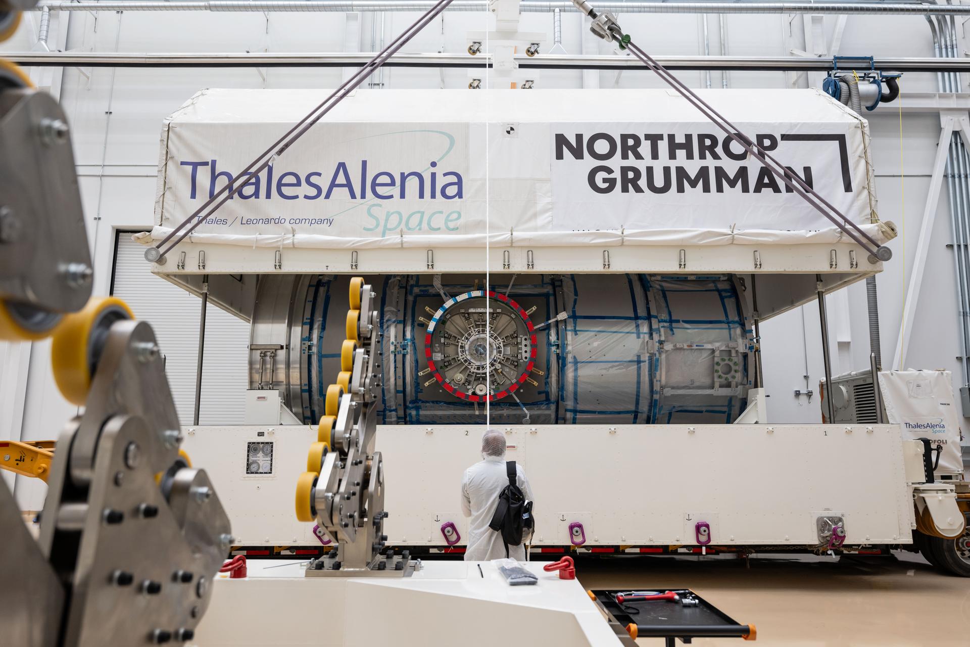 A technician in cleanroom attire stands in front of HALO’s transport container as the top half is being lifted off inside Northrop Grumman’s facility in Gilbert, Arizona. The cylindrical HALO module is partially visible inside the container. The container is labeled with Thales Alenia Space and Northrop Grumman logos. Equipment and hardware are visible in the foreground.