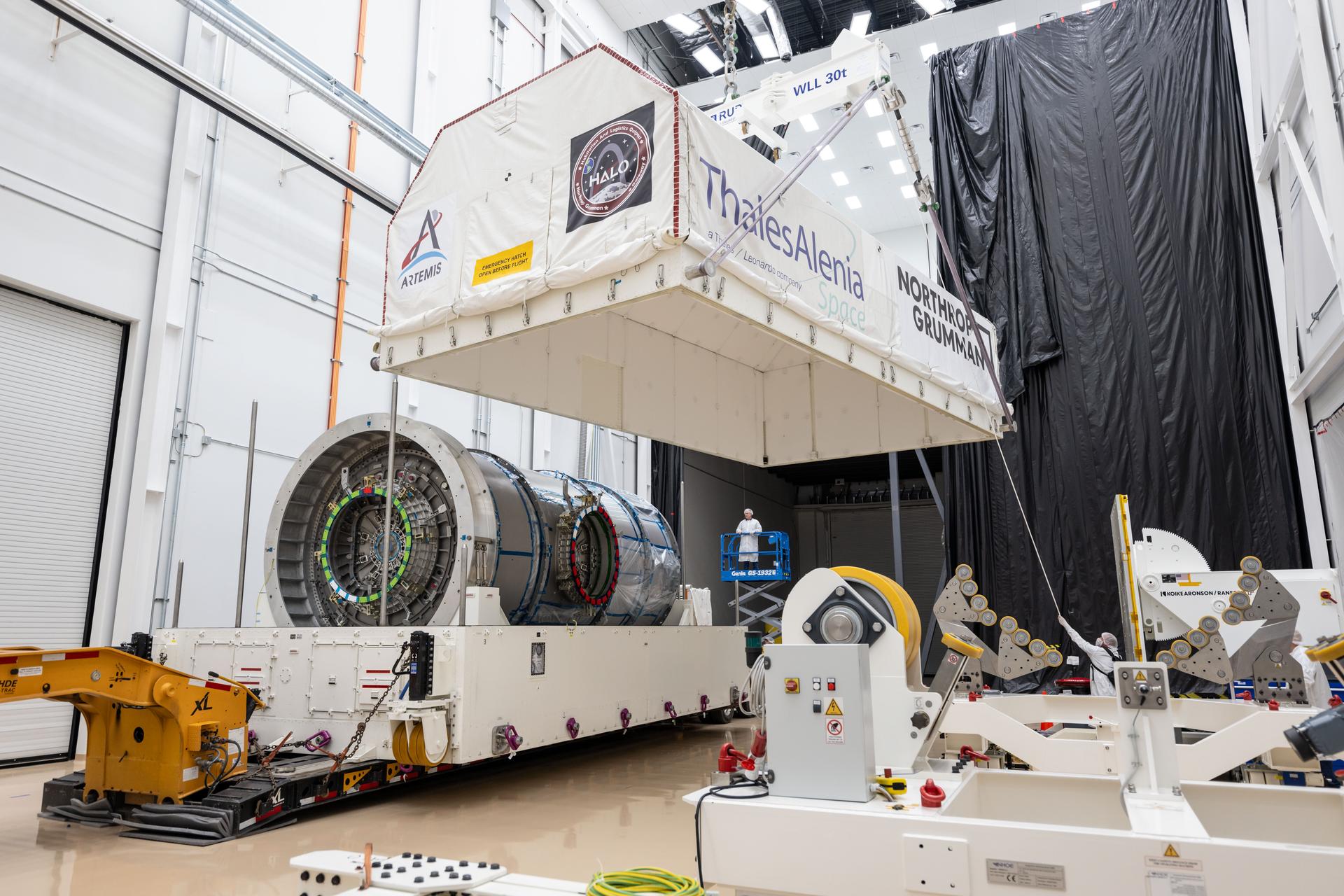 Inside Northrop Grumman’s facility in Gilbert, Arizona, the top half of the HALO module’s transport container is lifted away, revealing the metallic cylindrical structure beneath. The container is marked with logos for Artemis, HALO, Thales Alenia Space, and Northrop Grumman. A technician on a lift observes the operation.