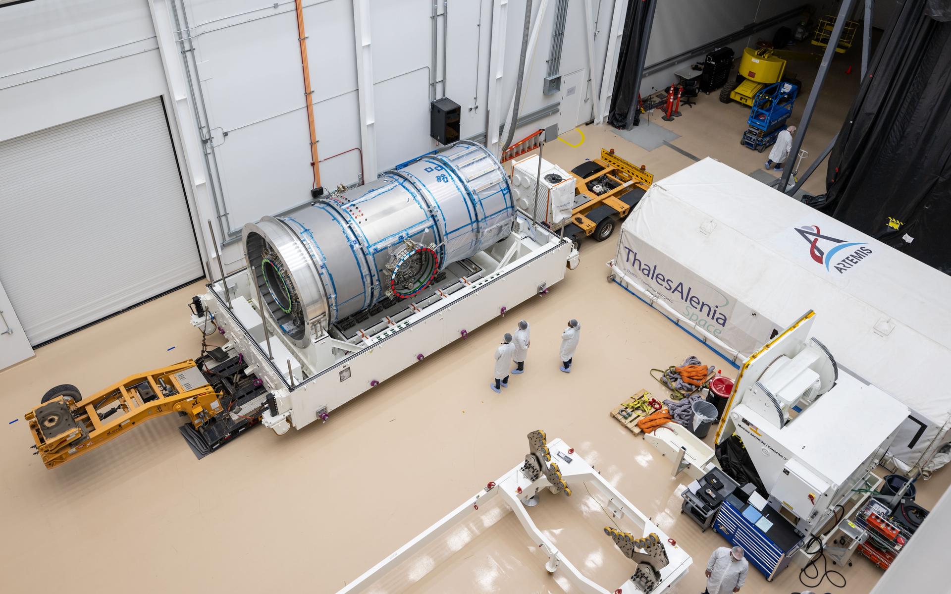 Overhead view inside Northrop Grumman’s facility in Gilbert, Arizona, showing Gateway's HALO module secured in a white support structure after removal of the top of its transport container. The metallic cylindrical module is surrounded by personnel in cleanroom garments. The opened shipping container, marked with Thales Alenia Space and Artemis logos, is positioned nearby along with handling equipment and tools.