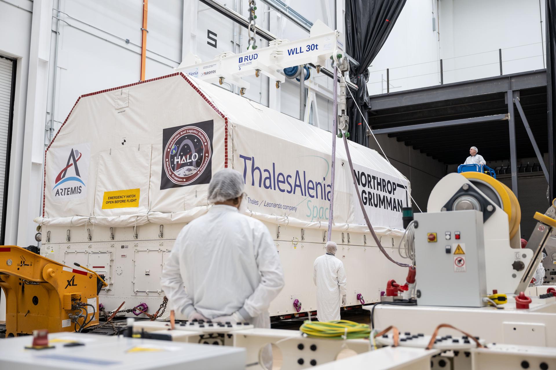 Personnel work around HALO’s transport container inside Northrop Grumman’s facility in Gilbert, Arizona. The white container is suspended from an overhead crane and features logos for Artemis, HALO, Thales Alenia Space, and Northrop Grumman. A technician on a lift observes from above while others prepare for unloading.
