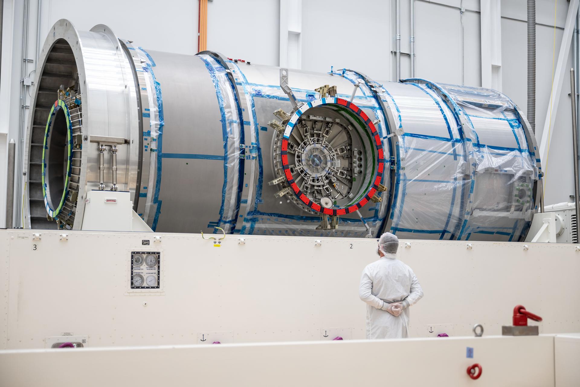A cleanroom technician stands with hands behind their back, observing Gateway’s HALO module inside Northrop Grumman’s facility in Gilbert, Arizona. The metallic cylindrical structure rests in its support cradle, partially wrapped in protective film, with visible docking ports and alignment markings.