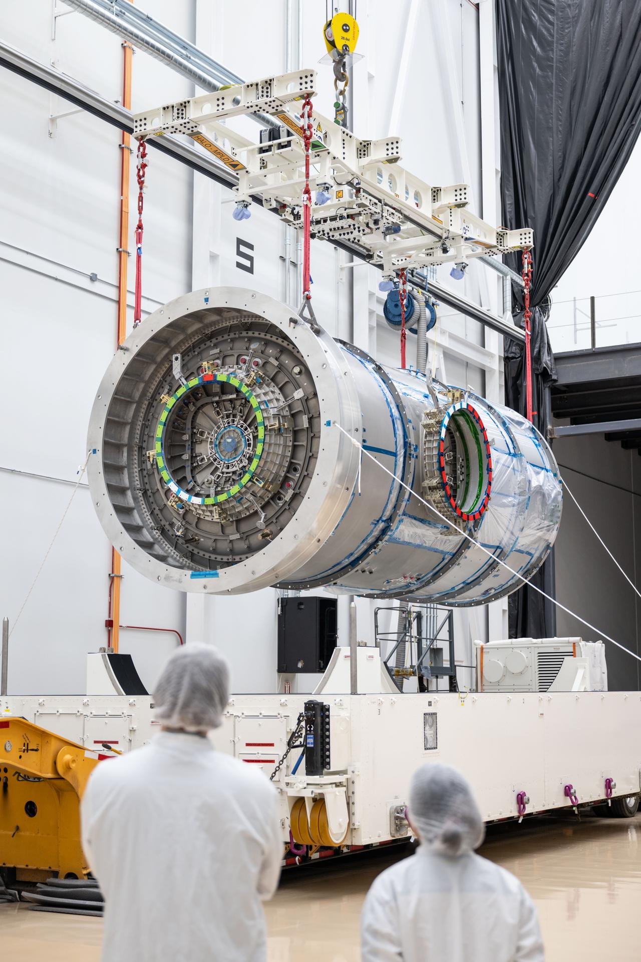 Two technicians observe as Gateway’s HALO module is suspended midair by a crane inside Northrop Grumman’s facility in Gilbert, Arizona. The cylindrical module, with visible interface ports and wrapping, is lifted from its transport container using a large white lifting fixture with red straps.