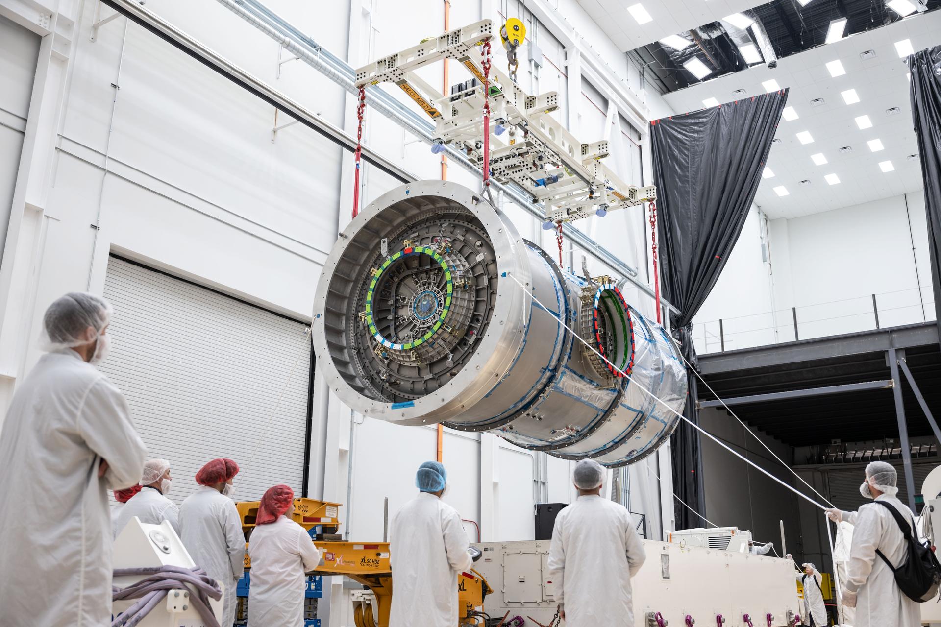 A group of technicians observe as Gateway’s HALO module is suspended midair by a crane inside Northrop Grumman’s facility in Gilbert, Arizona. The cylindrical metallic structure is lifted using red straps and a large white lifting fixture.