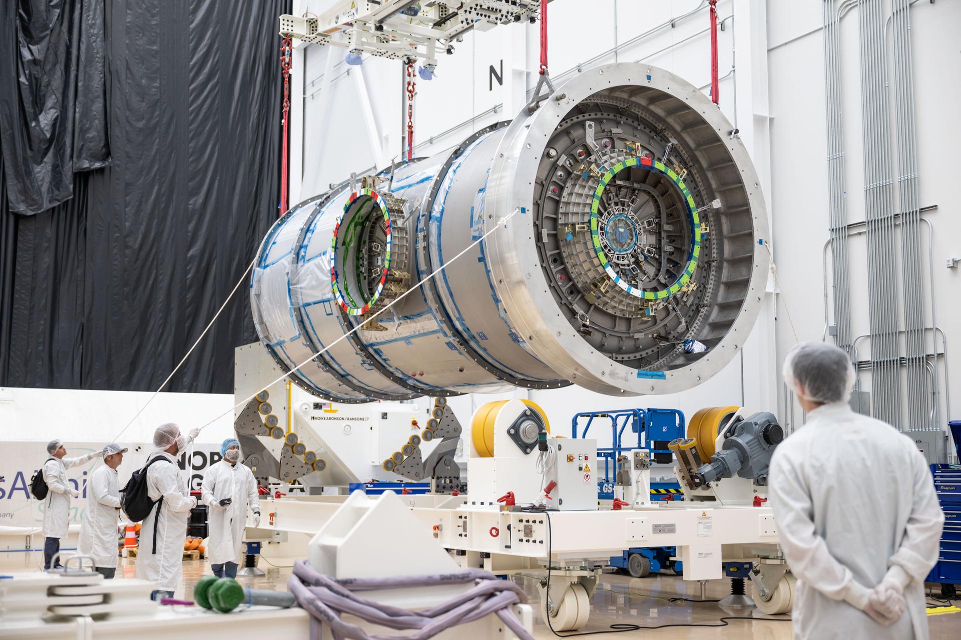 Inside Northrop Grumman’s facility in Gilbert, Arizona, Gateway’s HALO module is suspended in midair by a crane as technicians in cleanroom garments observe the operation. The cylindrical metallic module features docking interfaces with alignment markings. Surrounding the module is specialized handling equipment used to support its movement.