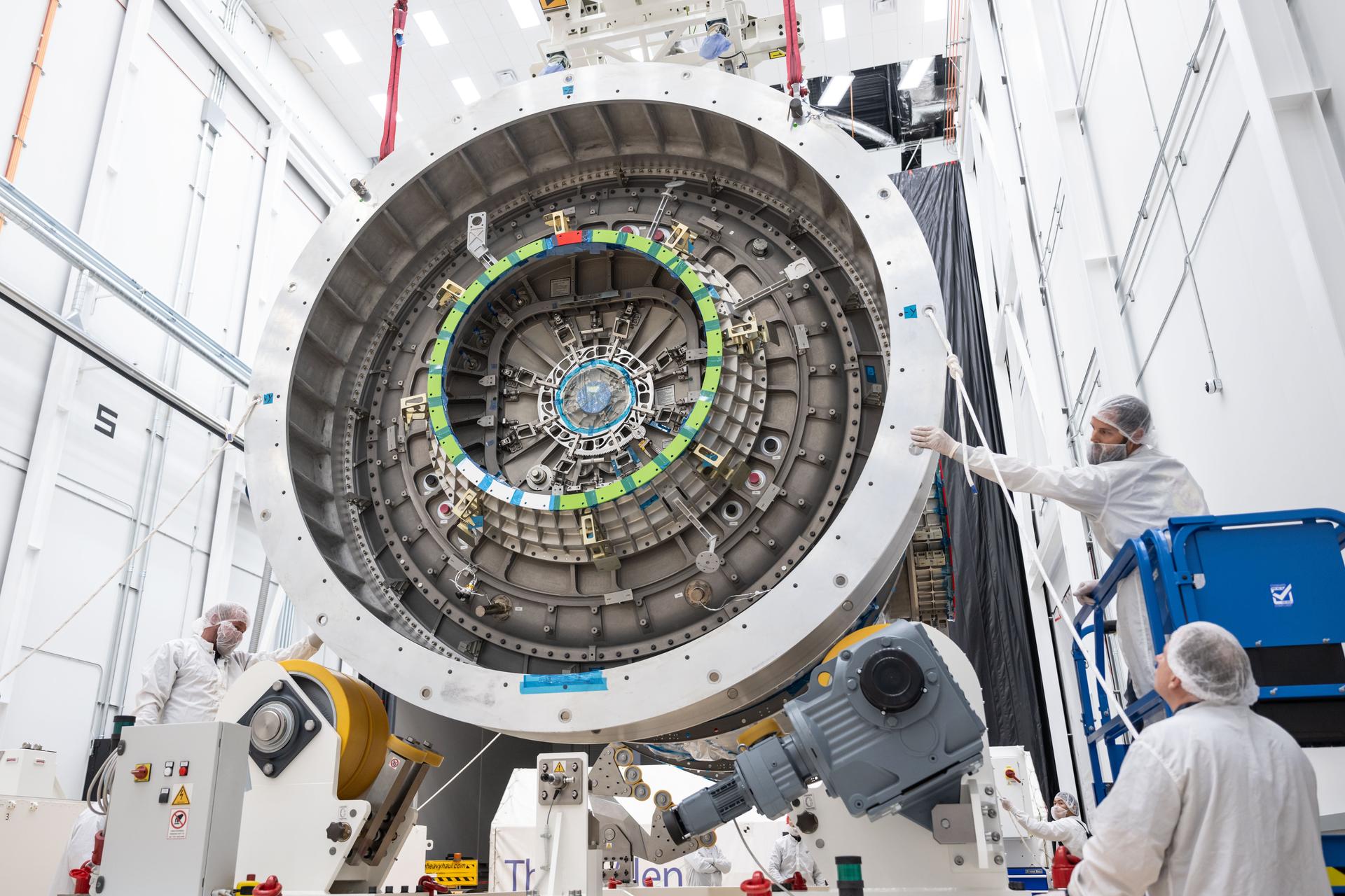 Technicians in cleanroom attire guide and inspect Gateway’s HALO module as it is suspended in midair inside Northrop Grumman’s facility in Gilbert, Arizona. The close-up view shows intricate interface details and alignment markers on the circular docking port. One technician stands on a lift while others remain on the ground, coordinating the module’s position.