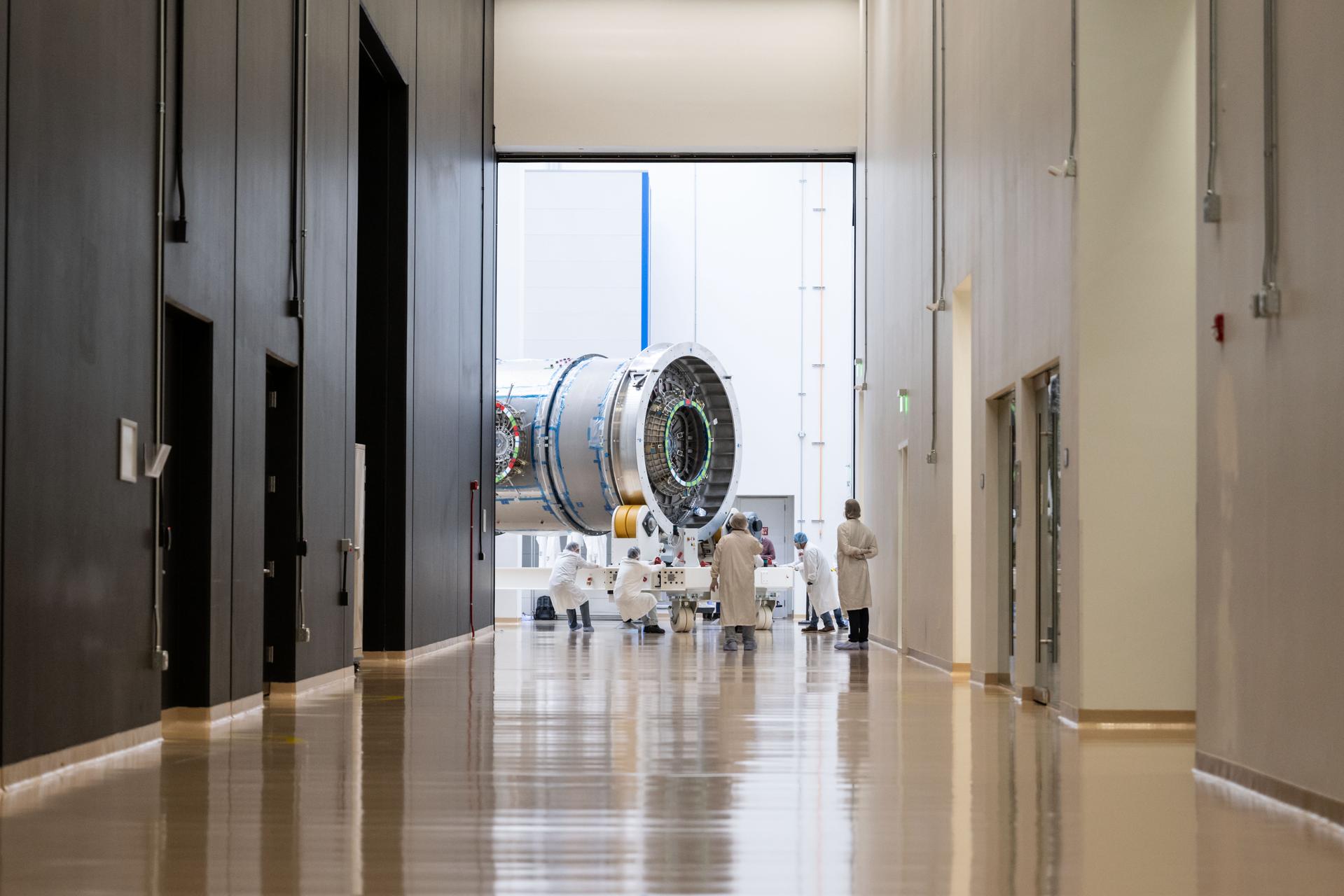 In a hallway, cleanroom technicians transport Gateway’s HALO module inside Northrop Grumman’s facility in Gilbert, Arizona. The cylindrical metallic module is centered in a doorway at the end of the corridor as teams work to roll it down the hall.