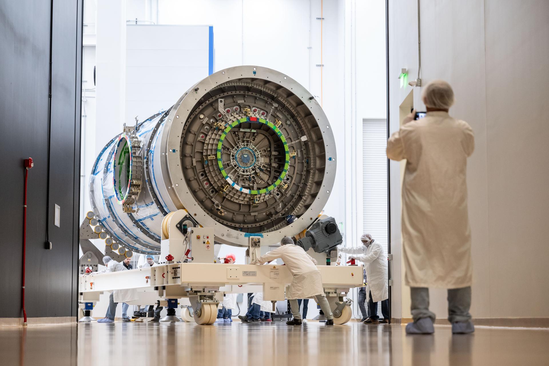 A cleanroom technician takes a photo as a team moves Gateway’s HALO module down a hallway inside Northrop Grumman’s facility in Gilbert, Arizona. The cylindrical metallic module, with visible alignment markings and docking port, is supported on a white wheeled platform.