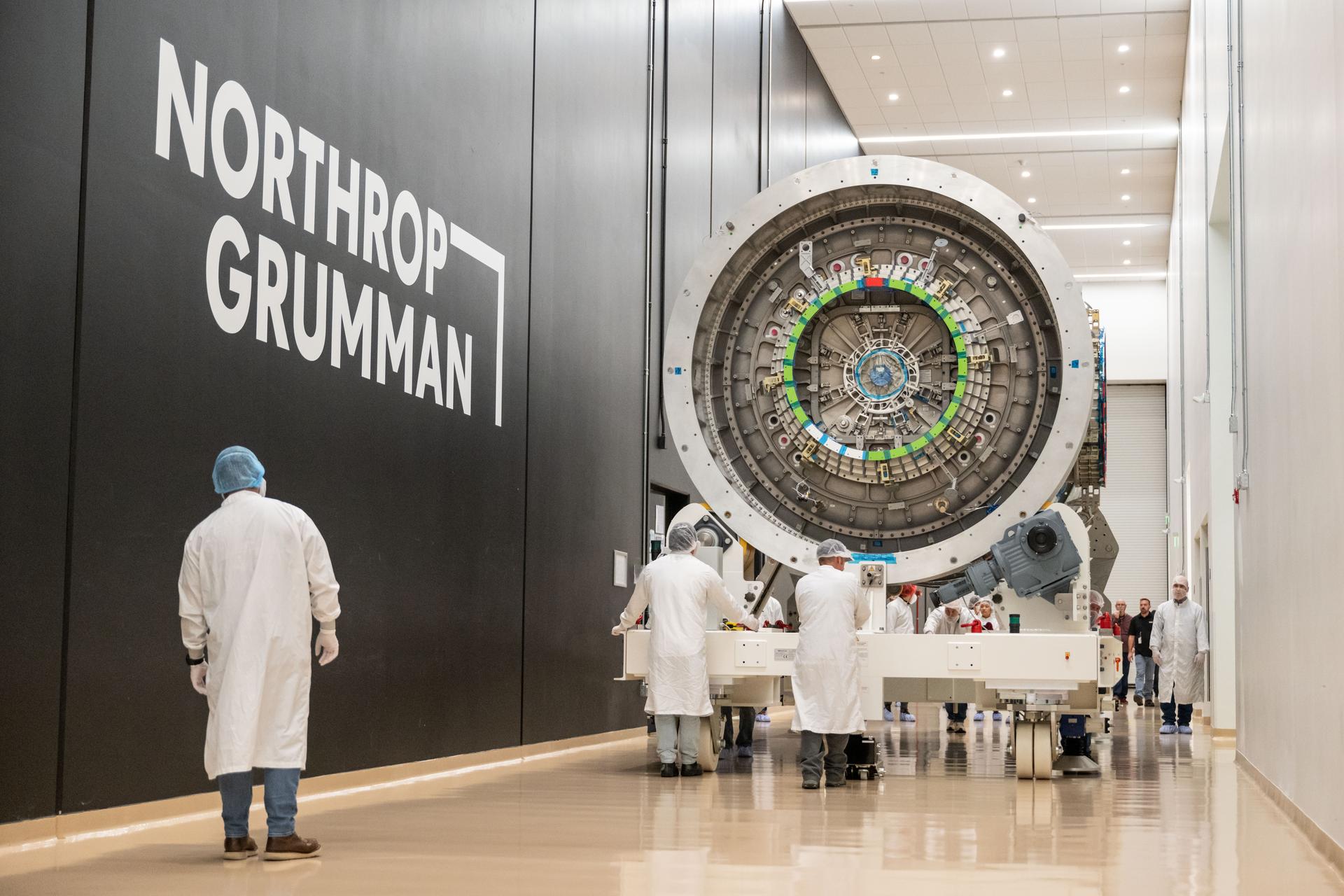 team moves Gateway’s HALO module down a hallway inside Northrop Grumman’s facility in Gilbert, Arizona. The cylindrical metallic module, with visible alignment markings and docking port, is supported on a white wheeled platform.