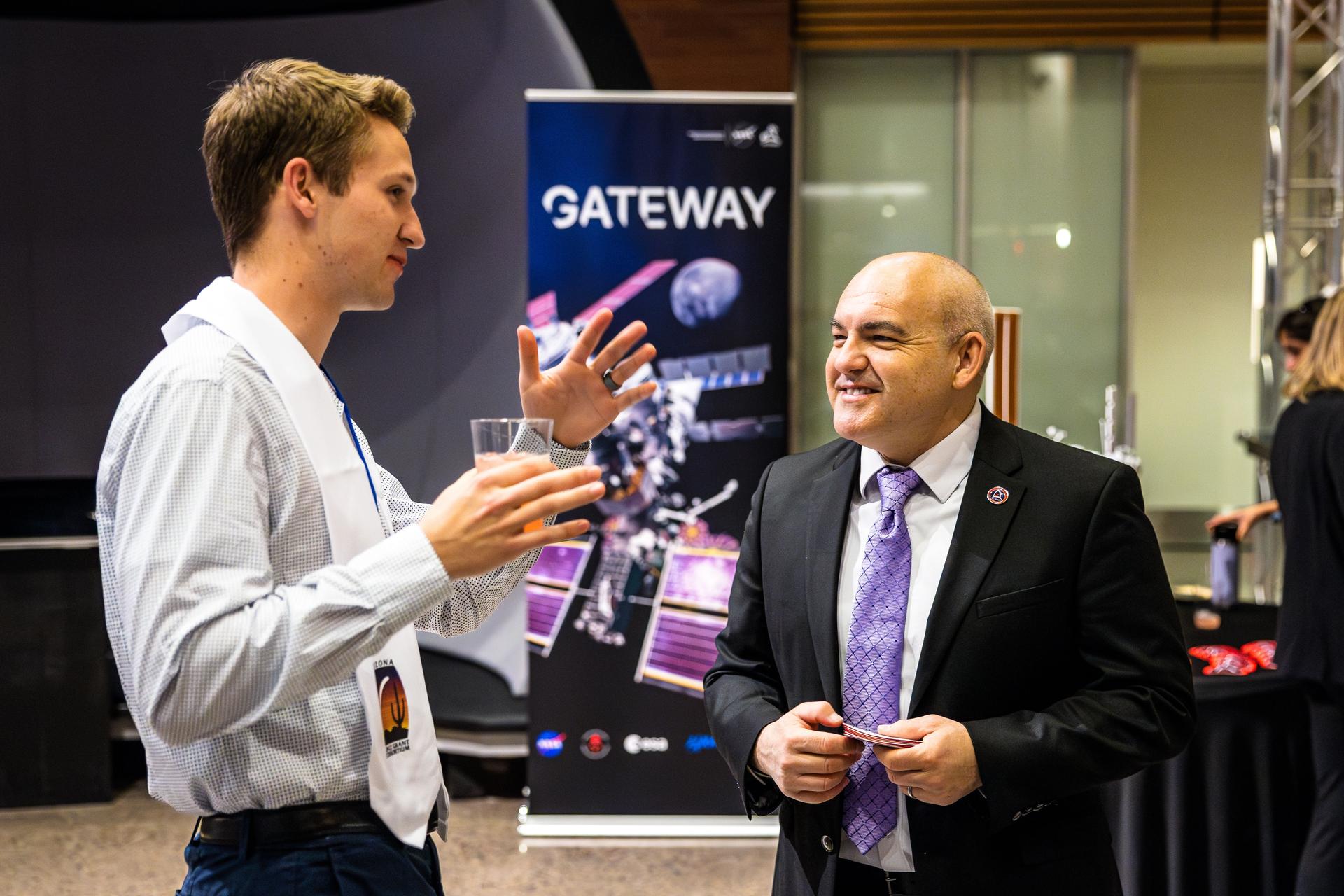 Carlos Garcia-Galan, wearing a black suit and purple tie, smiles while speaking with a college student holding a glass of soda during the 2025 Arizona NASA Space Grant Consortium Statewide Student Research Symposium at Arizona State University. A Gateway program banner is visible in the background.