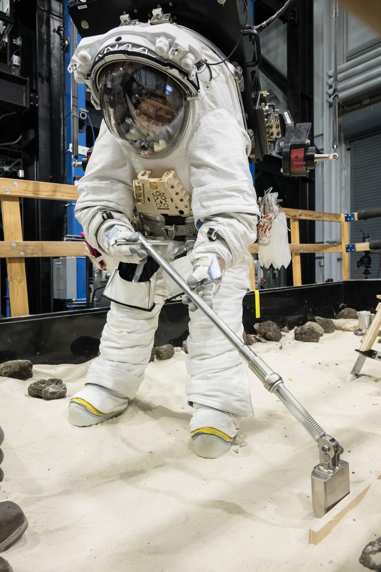 A NASA crew member practices using lunar tools to collect geology samples at NASA’s Johnson Space Center during an elevated suit pressure test where teams evaluate how well crew perform tasks in different suit pressure levels while wearing the Artemis III lunar spacesuit developed by Axiom Space called the AxEMU (Axiom Extravehicular Mobility Unit).