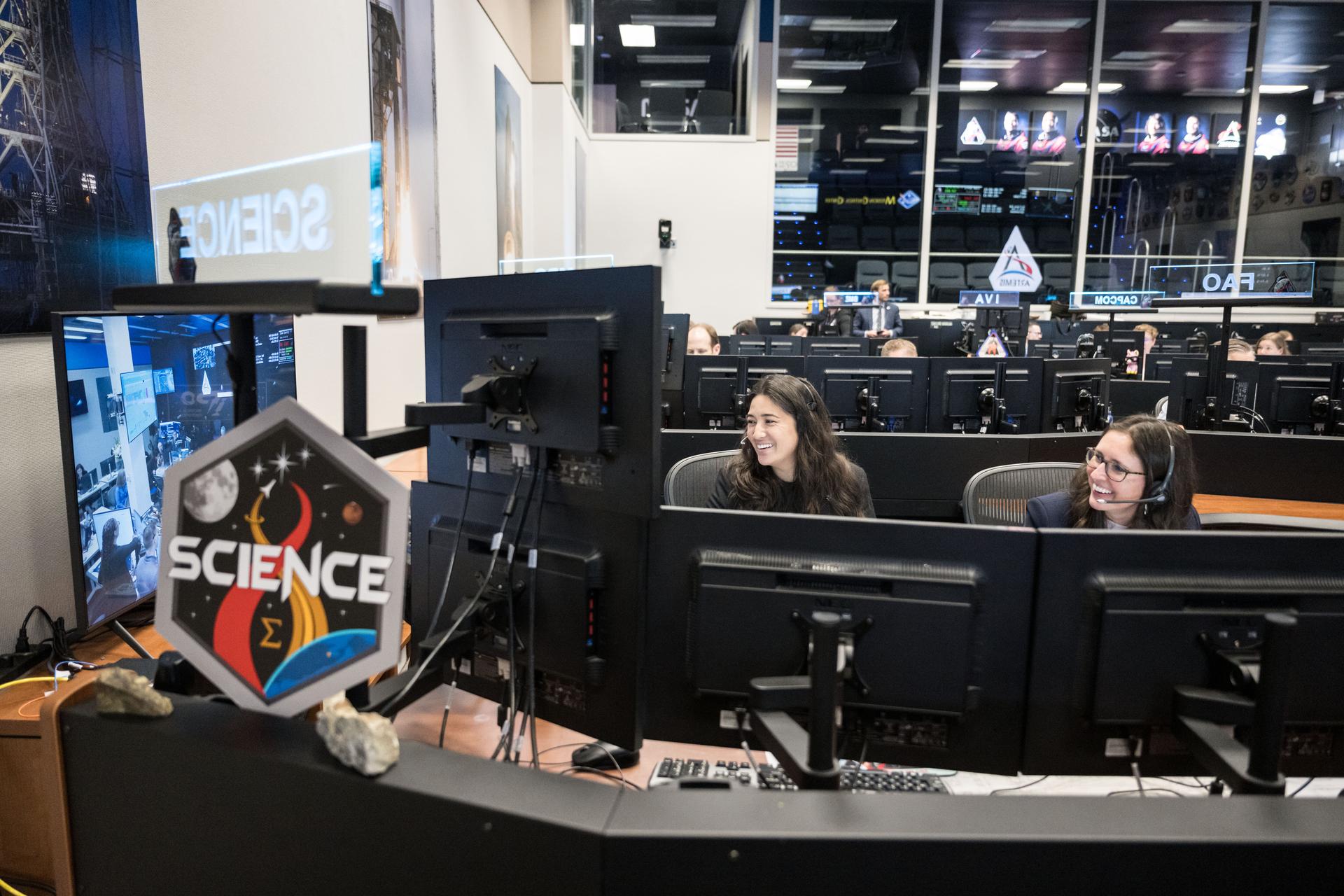 Artemis science officers, from left, Angela Garcia and Kelsey Young, watch the lunar science team celebrating in the Science Evaluation Room (SER) as they hear lunar observations from the Artemis II crew. The science team has spent years training the astronauts in geology and observation, both in the classroom and in the field. They also built the lunar targeting plan that, like a spacewalk plan, provides strong, detailed observation guidance, plus flexibility for the crew to make decisions based on what they’re seeing and experiencing in real time. The science team had many moments of celebration during the lunar flyby as the astronauts took pictures of the Moon and provided verbal descriptions of what they were seeing. This type of information reveals the geologic history of an area and will be critical to collect when future Artemis astronauts explore the Moon's surface. Credits: NASA/Robert Markowitz