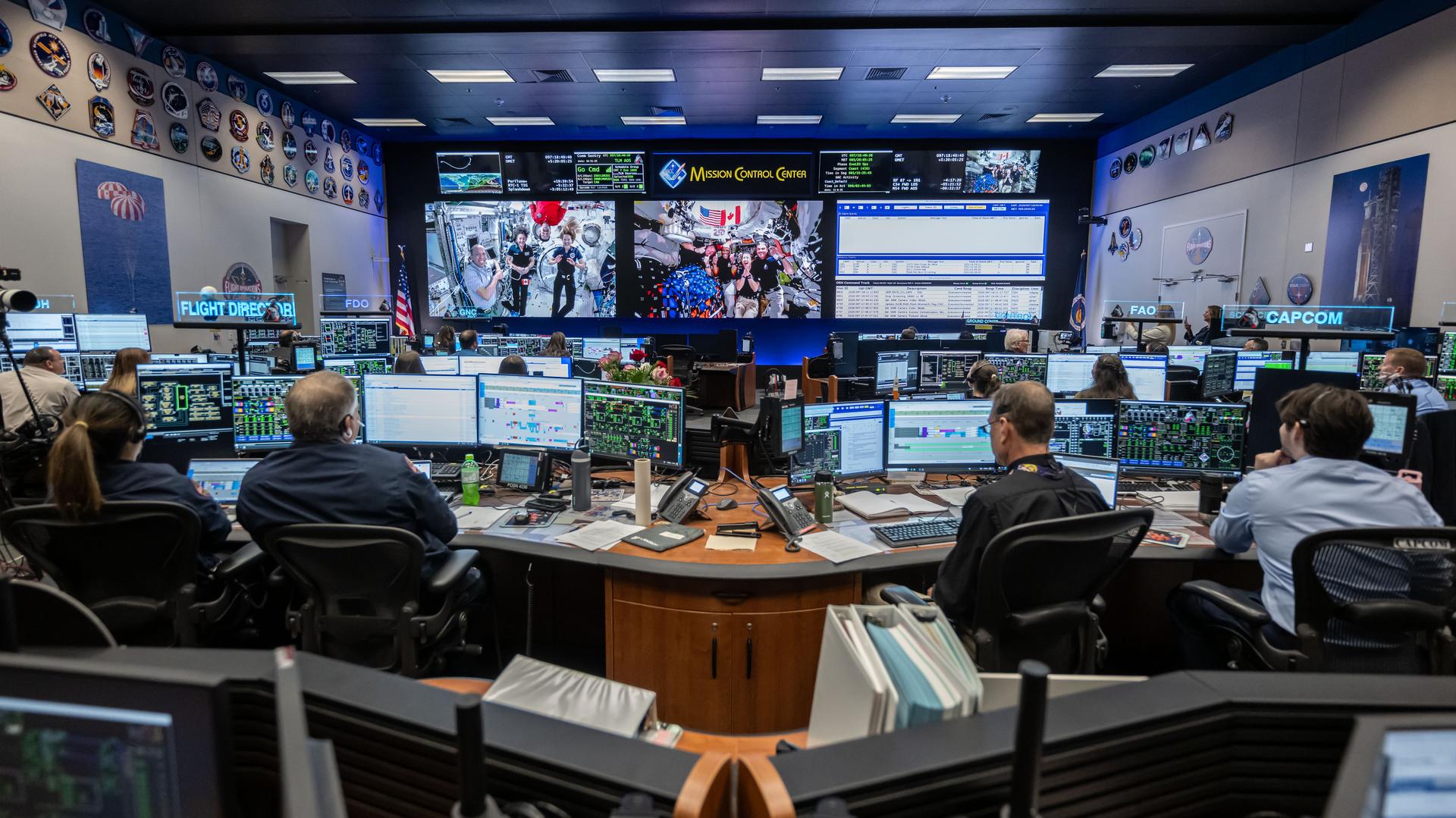 Two screens at the front of Mission Control show three astronauts floating inside the International Space Station on the left and four astronauts floating inside the Orion space craft. A third screen shows writing. In the foreground, flight controllers watch from behind computer screens at their desks.