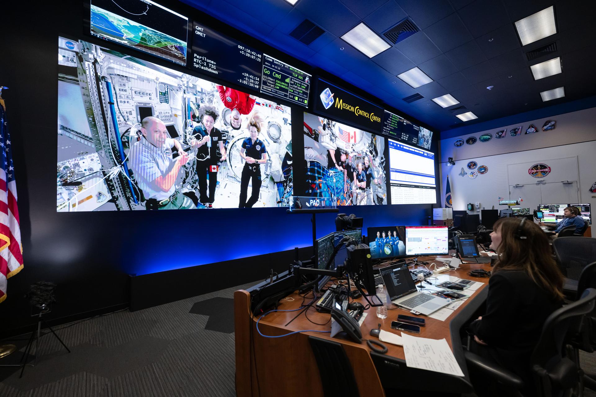 Two screens at the front of Mission Control show three astronauts floating at the International Space Station (left) and four astronauts floating in the Orion spacecraft (right). Flight controllers watch from behind computer screens at their desks.