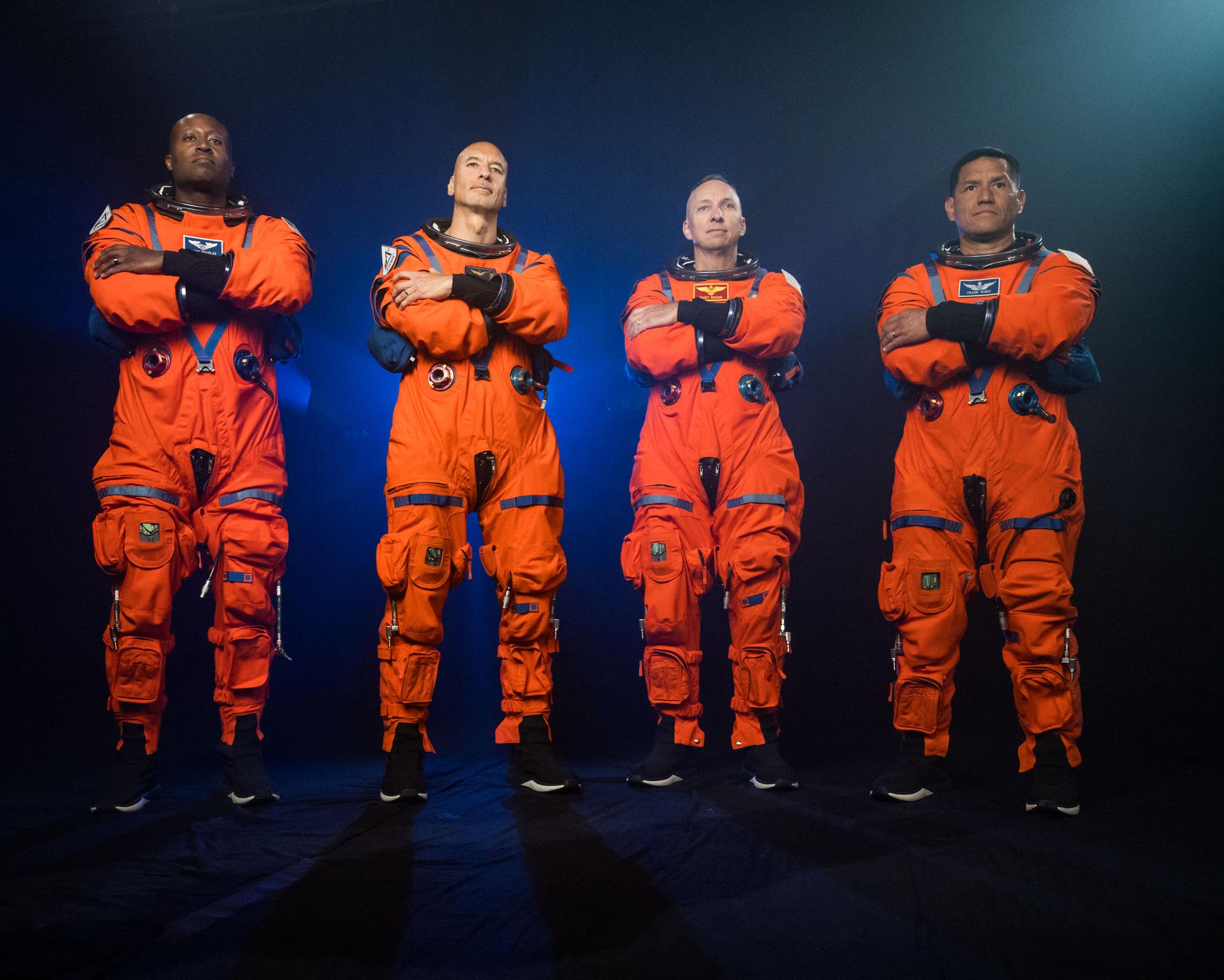Four astronauts are pictured wearing orange Orion Crew Survival System suits. The photo is illuminated with blue lights in the background. All four astronauts are standing next to each other with their arms crossed, looking away from the camera. 