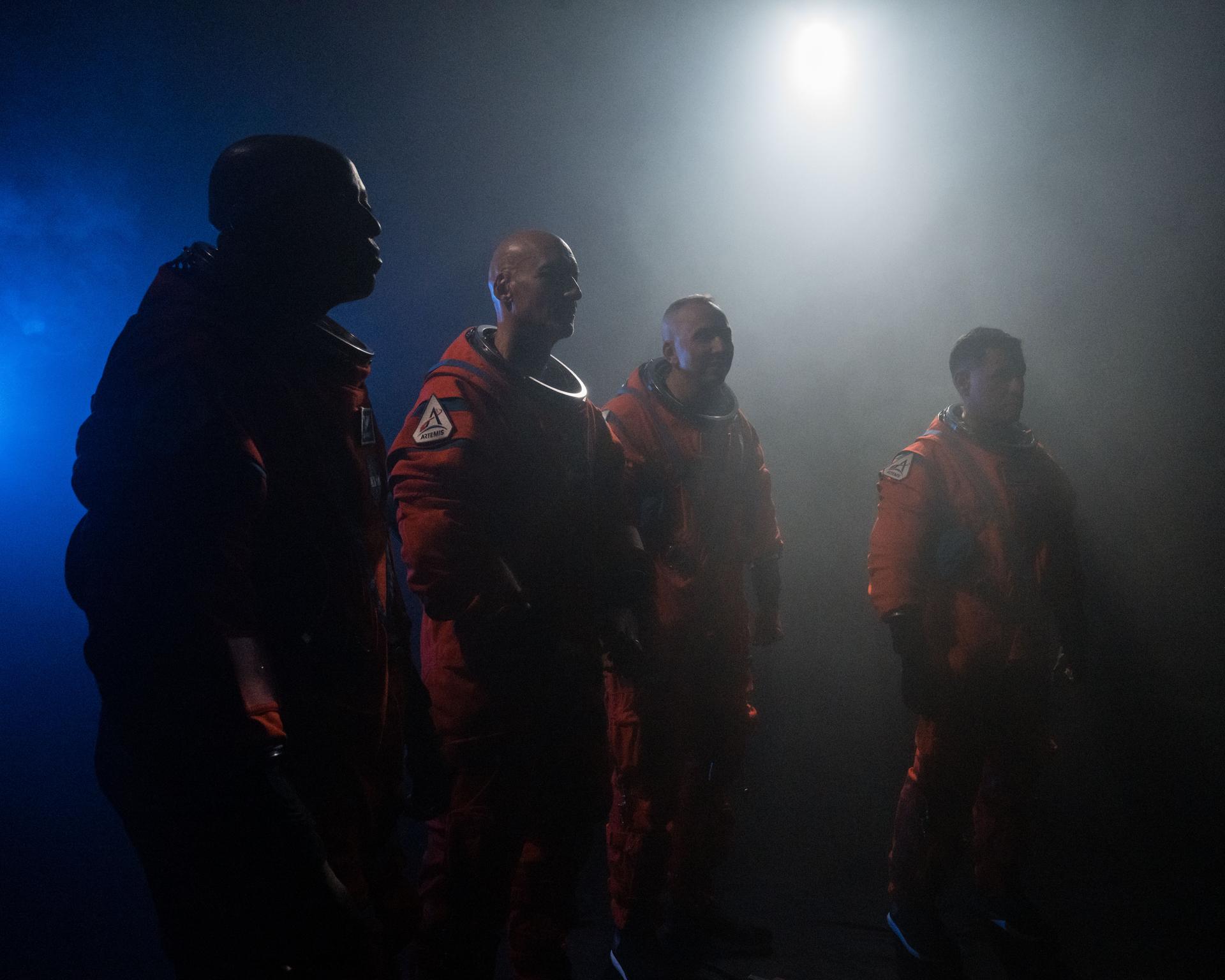 Four astronauts are pictured wearing orange Orion Crew Survival System suits. The photo is dark with a bright white light illuminating the far upper-right side with a blue light shining in the far left side. The astronauts are turned away from the camera with fog partially obscuring the image.