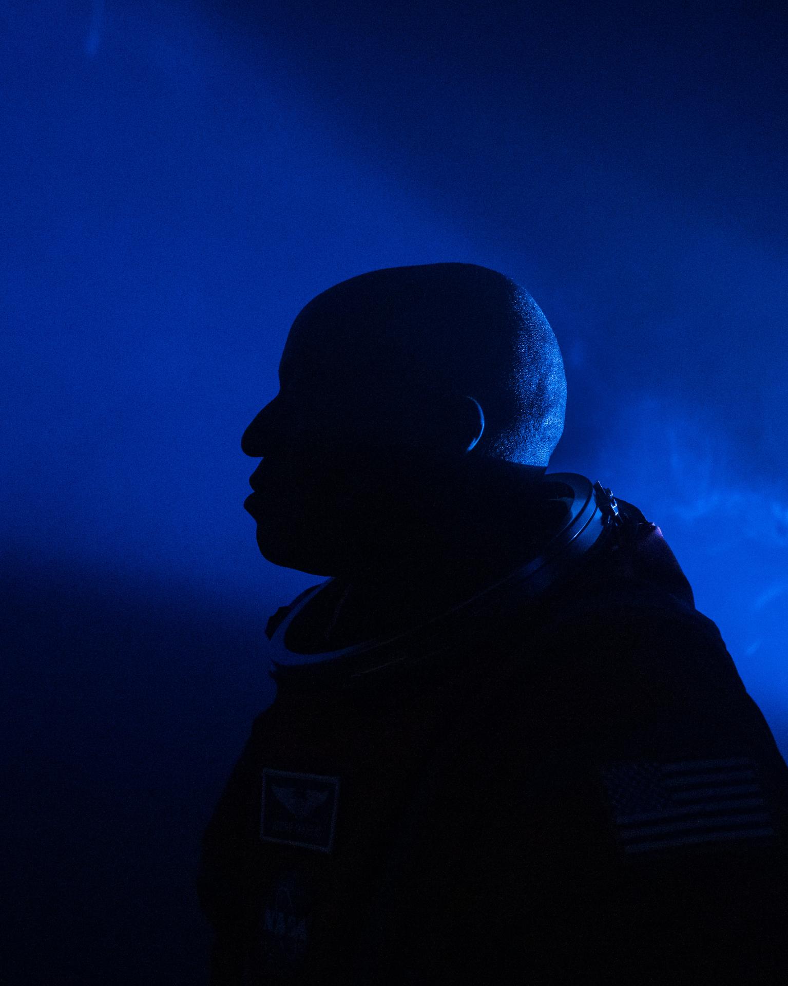 A side-profile, close-up silhouette of NASA astronaut Andre Douglas is pictured with blue lights and fog illuminating the background. He is pictured in an Orion Crew Survival System suit without the helmet.