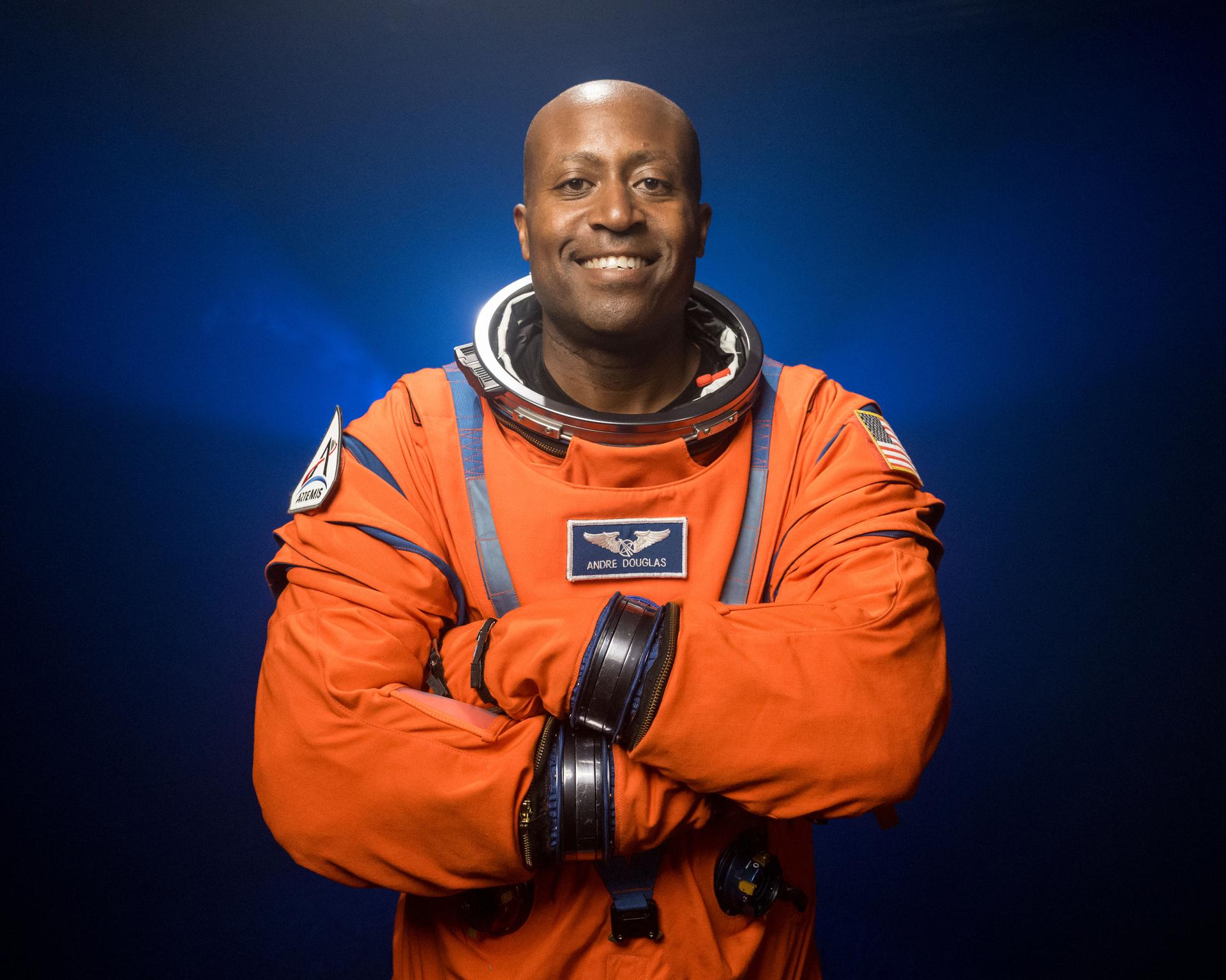 NASA astronaut Andre Douglas is pictured wearing an orange Orion Crew Survival System suit in a close-up shot. He is standing in front of a dark backdrop, illuminated with blue lights, with his arms crossed in front of his chest.