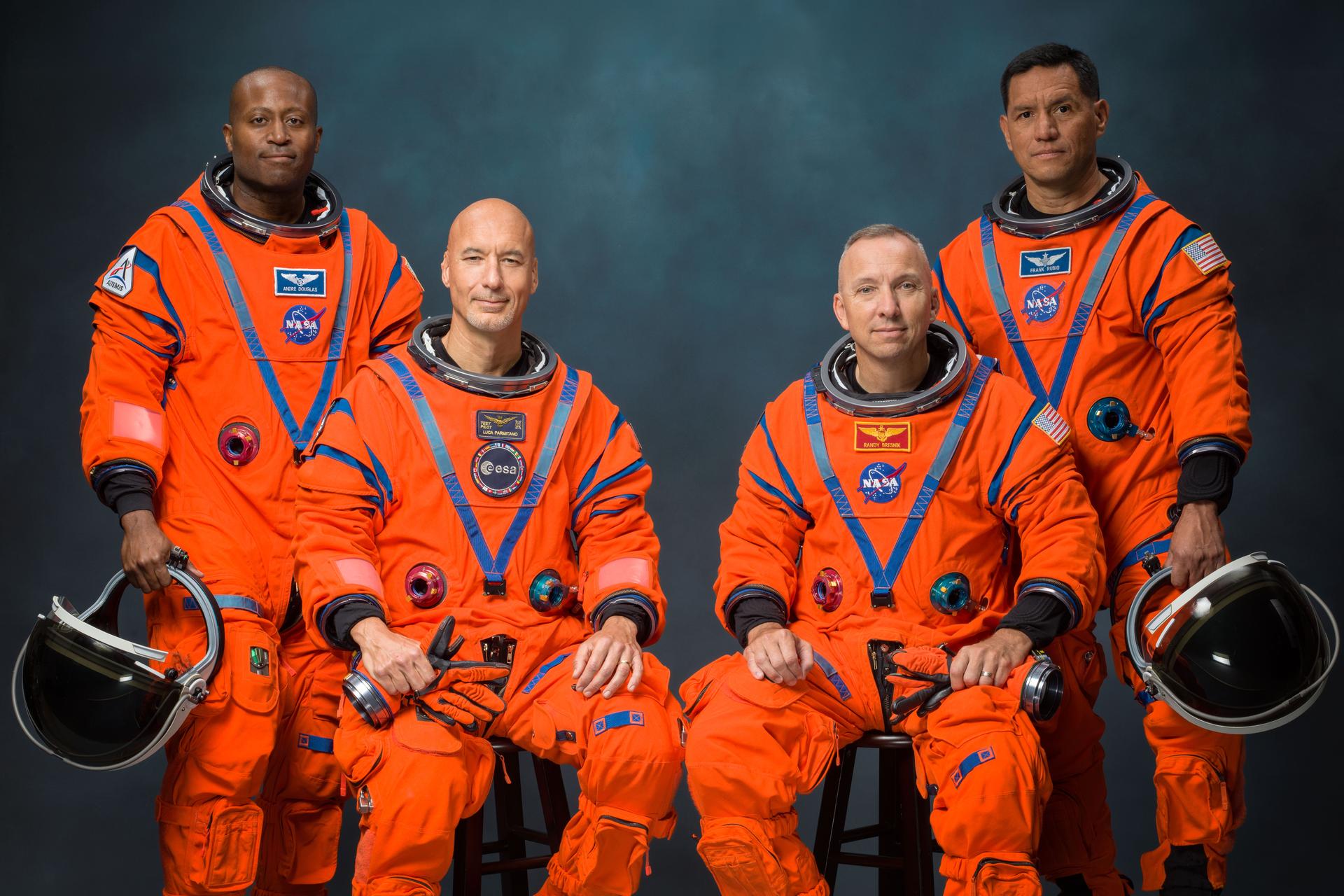 Four astronauts are pictured in orange Orion Crew Survival System suits. The astronauts on the far left and far right of the image are standing, holding their helmets. In between them, are the other two astronauts, who are seated and holding their gloves.